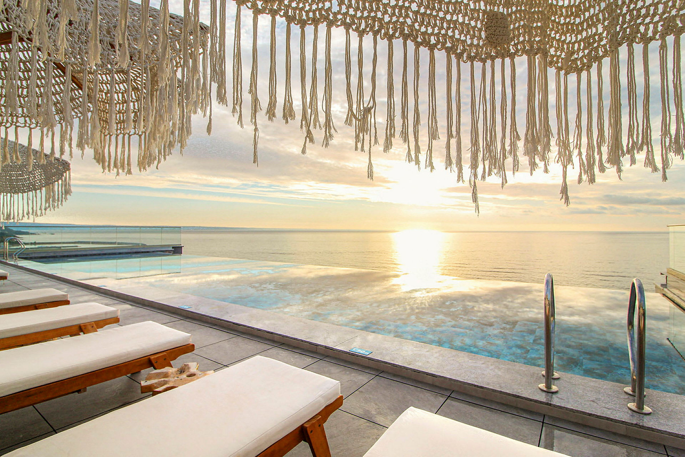 Solnedgång vid en infinitypool med solstolar i förgrunden och hängande dekorationer ovanför. (AI-genererad text)