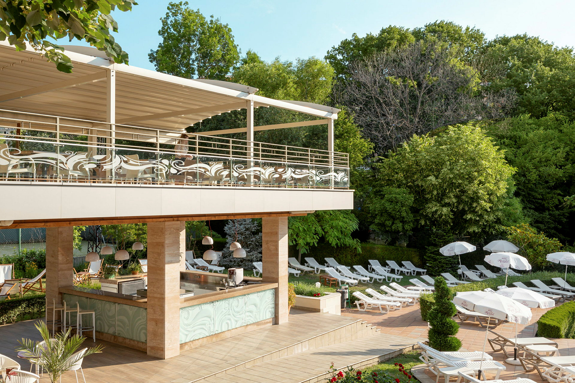 En utomhuspoolområde med solstolar, parasoller och en bar under ett övertäckt terrassområde. Området är omgivet av gröna träd. (AI-genererad text)