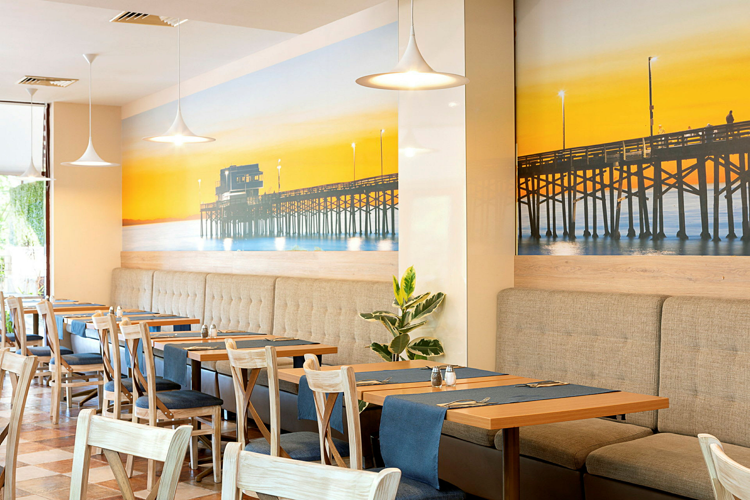 En restaurang med inredning som inkluderar bord och stolar, samt en väggdekoration föreställande en brygga vid solnedgången. (AI-genererad text)