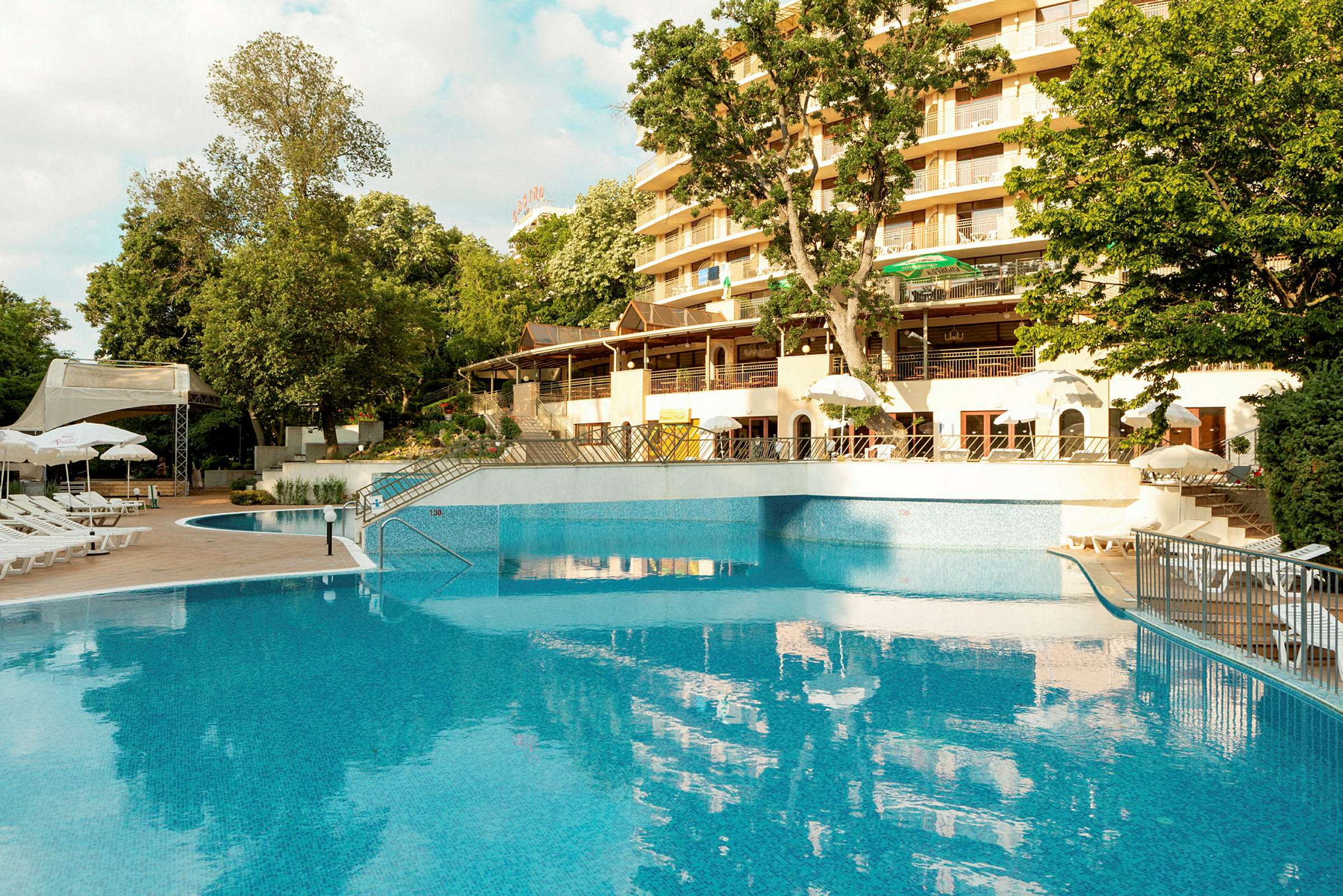 Bild av en pool vid ett hotell med solstolar och parasoller samt grönska och träd i bakgrunden. (AI-genererad text)