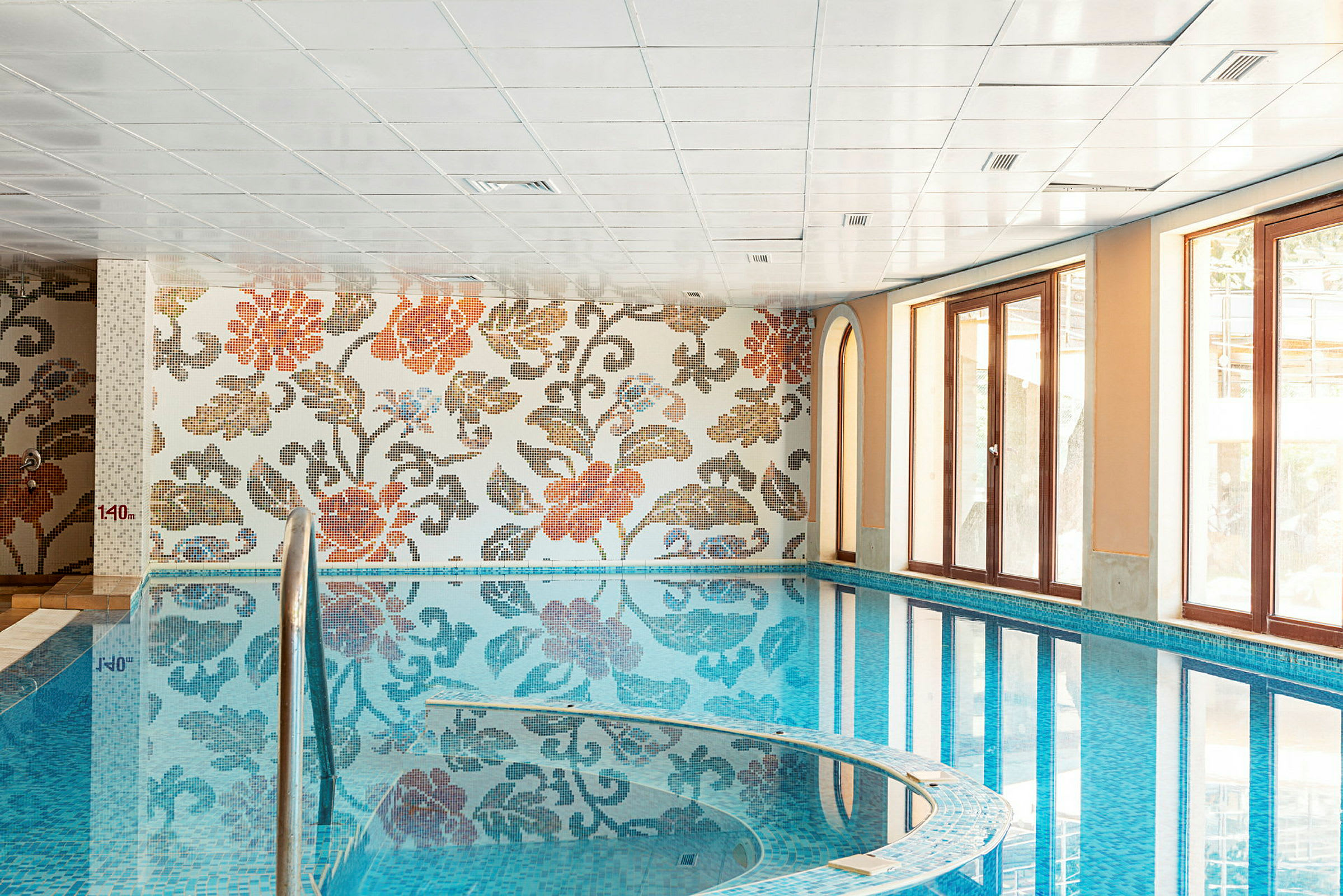 Inomhuspool med blommig mosaikvägg och stora fönster. (AI-genererad text)