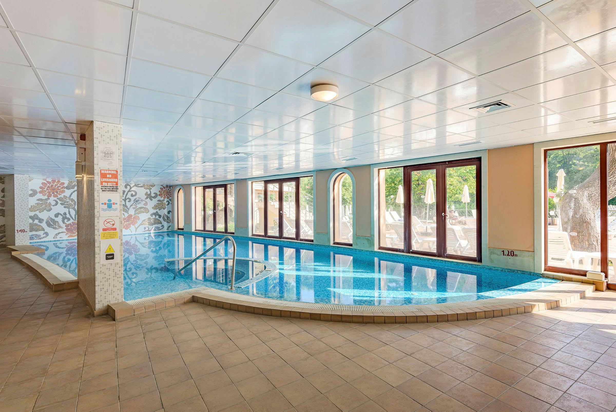 Inomhuspool med stora fönster mot en solig terrass. (AI-genererad text)