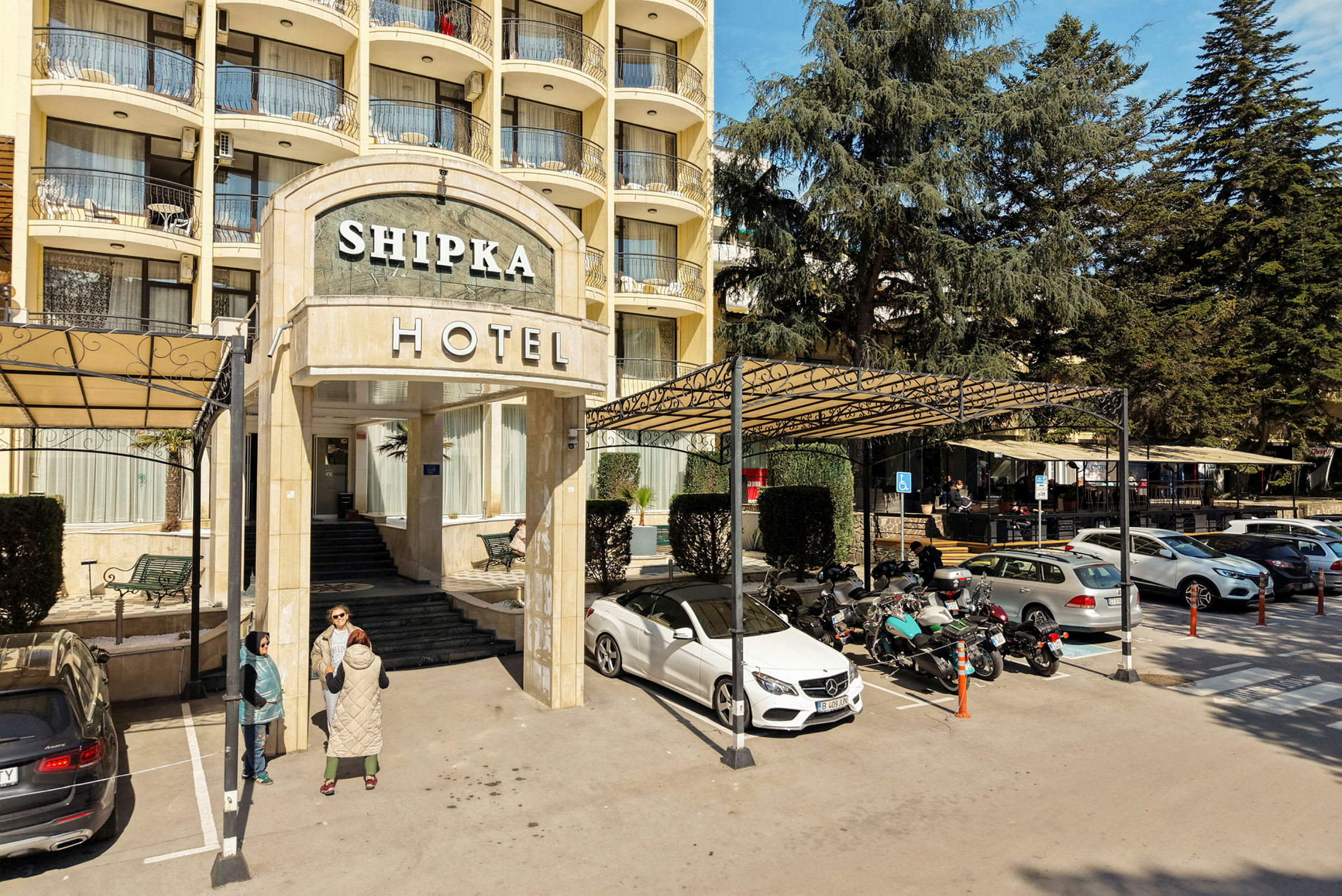 Hotellbyggnadens entré med trappa och skylt, omgivet av parkering med bilar och motorcyklar. (AI-genererad text)