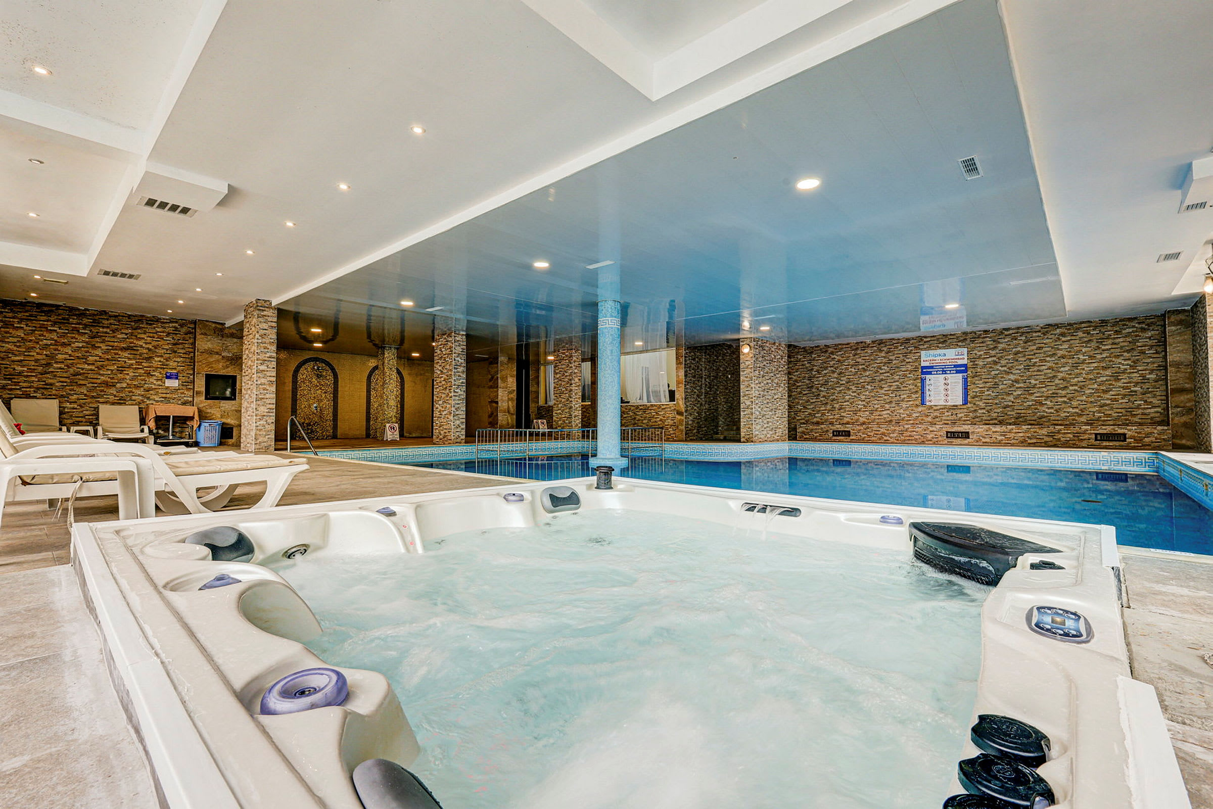 En inomhuspool med närliggande bubbelpool i ett utrymme med stenklädda väggar och takbelysning. (AI-genererad text)