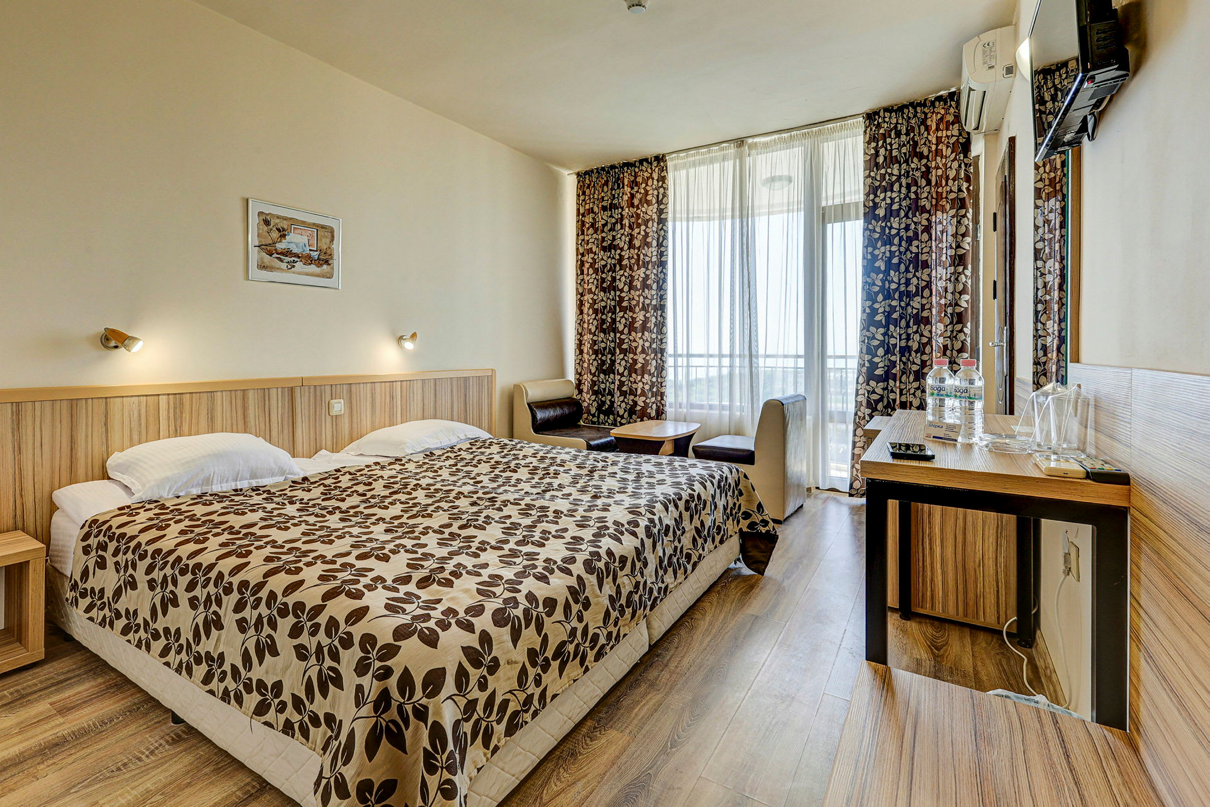 Ett hotellrum med en dubbelsäng, ett skrivbord med spegel och en sittgrupp vid ett fönster med gardiner. (AI-genererad text)