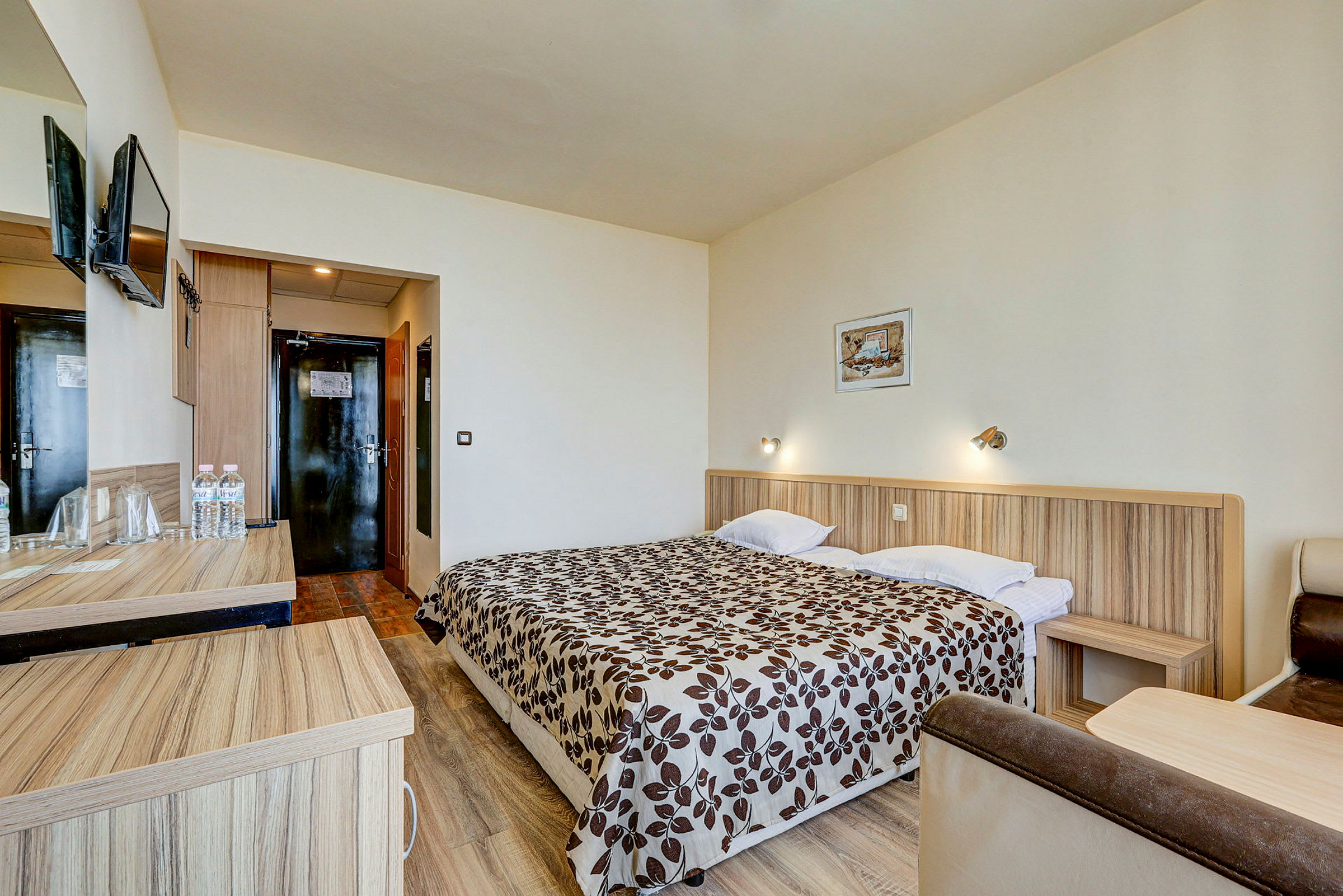 Ett hotellrum med en dubbelsäng med mönstrat täcke, ett skrivbord med vattenflaskor och en väggmonterad TV. (AI-genererad text)