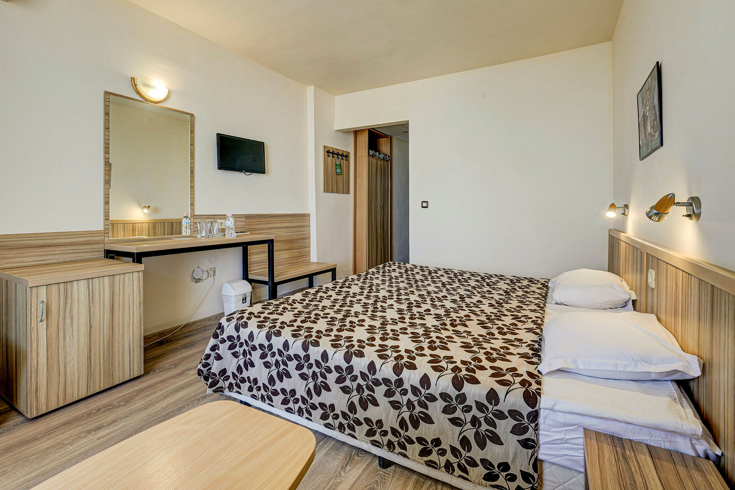 Ett hotellrum med en dubbelsäng med mönstrat överkast, sänglampor på väggen, ett skrivbord med spegel och stolar samt en TV på väggen. (AI-genererad text)