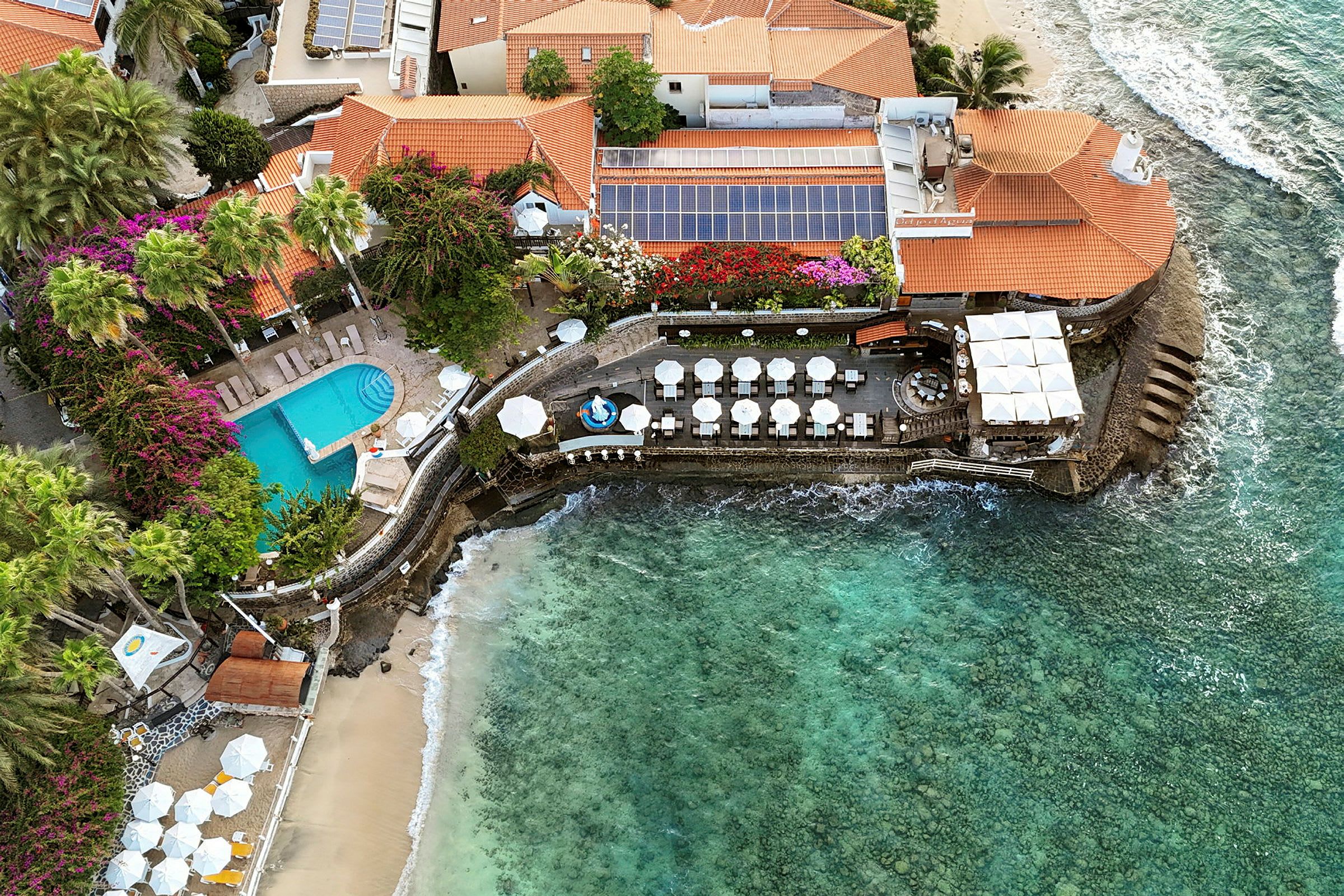 Flygfoto av en byggnad vid havet med pool och uteplatser. (AI-genererad text)