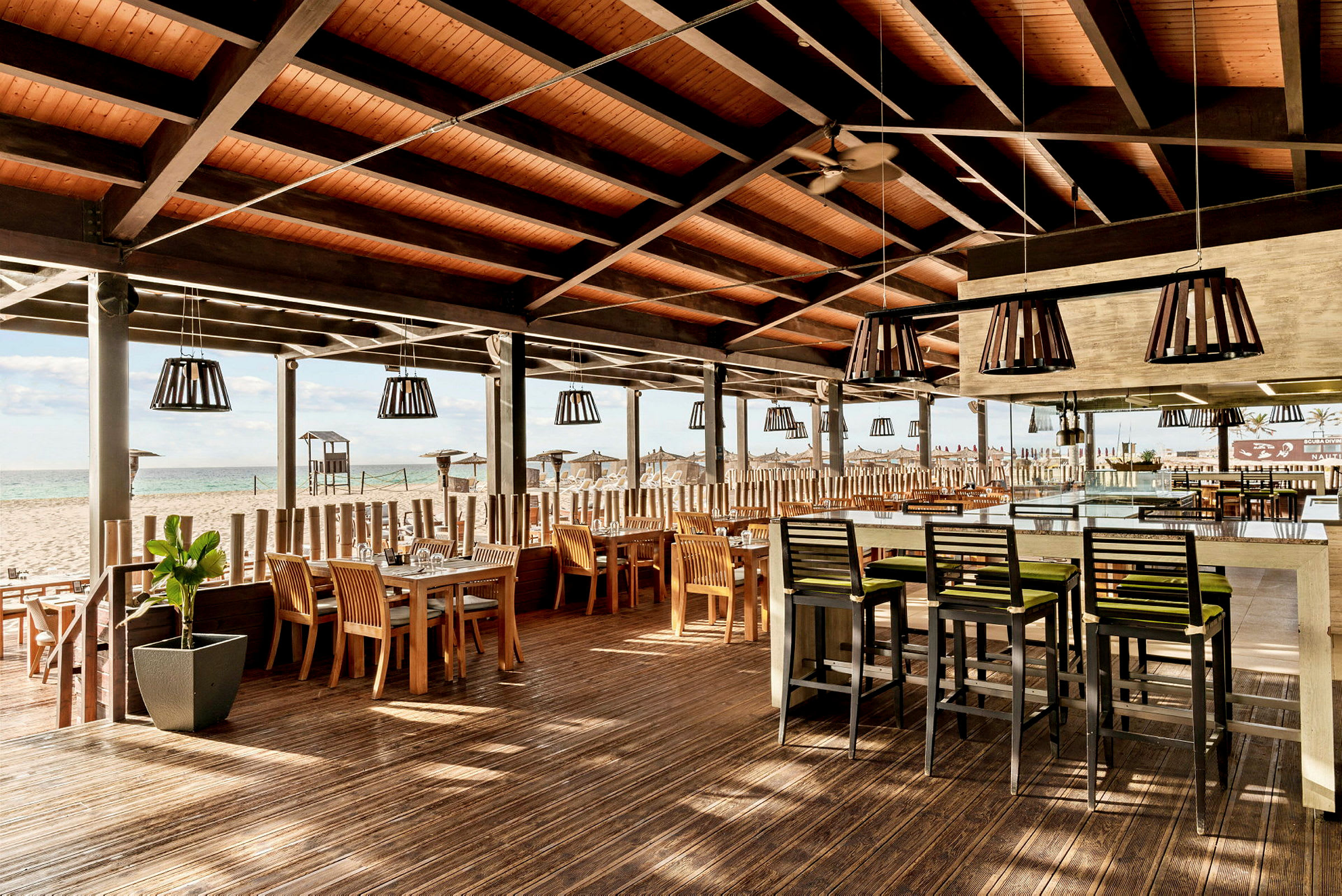 Utomhusrestaurang med tak, trästolar och bord vid en strand. (AI-genererad text)