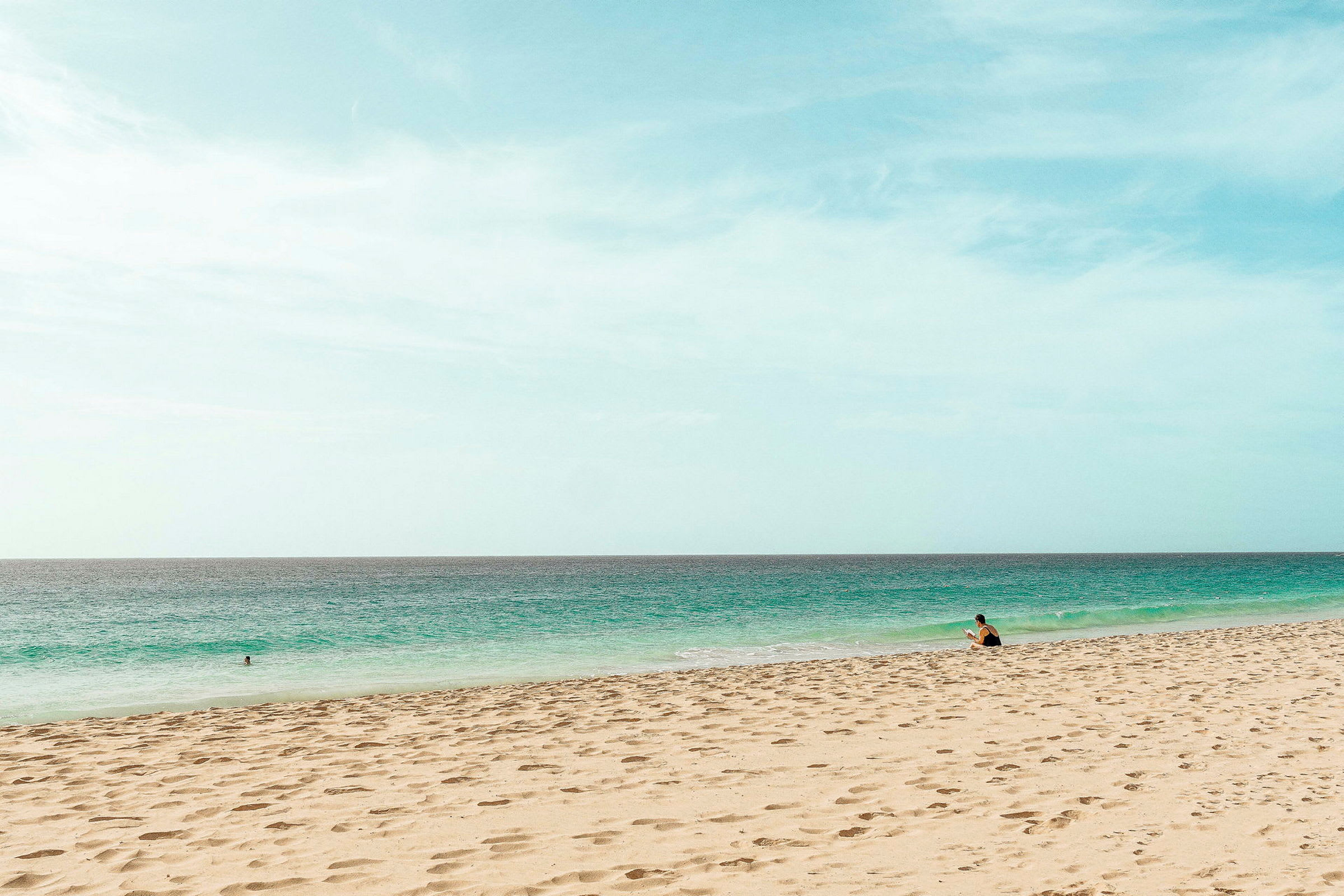 En strand med sand och hav, med en person som sitter nära vattenbrynet. (AI-genererad text)