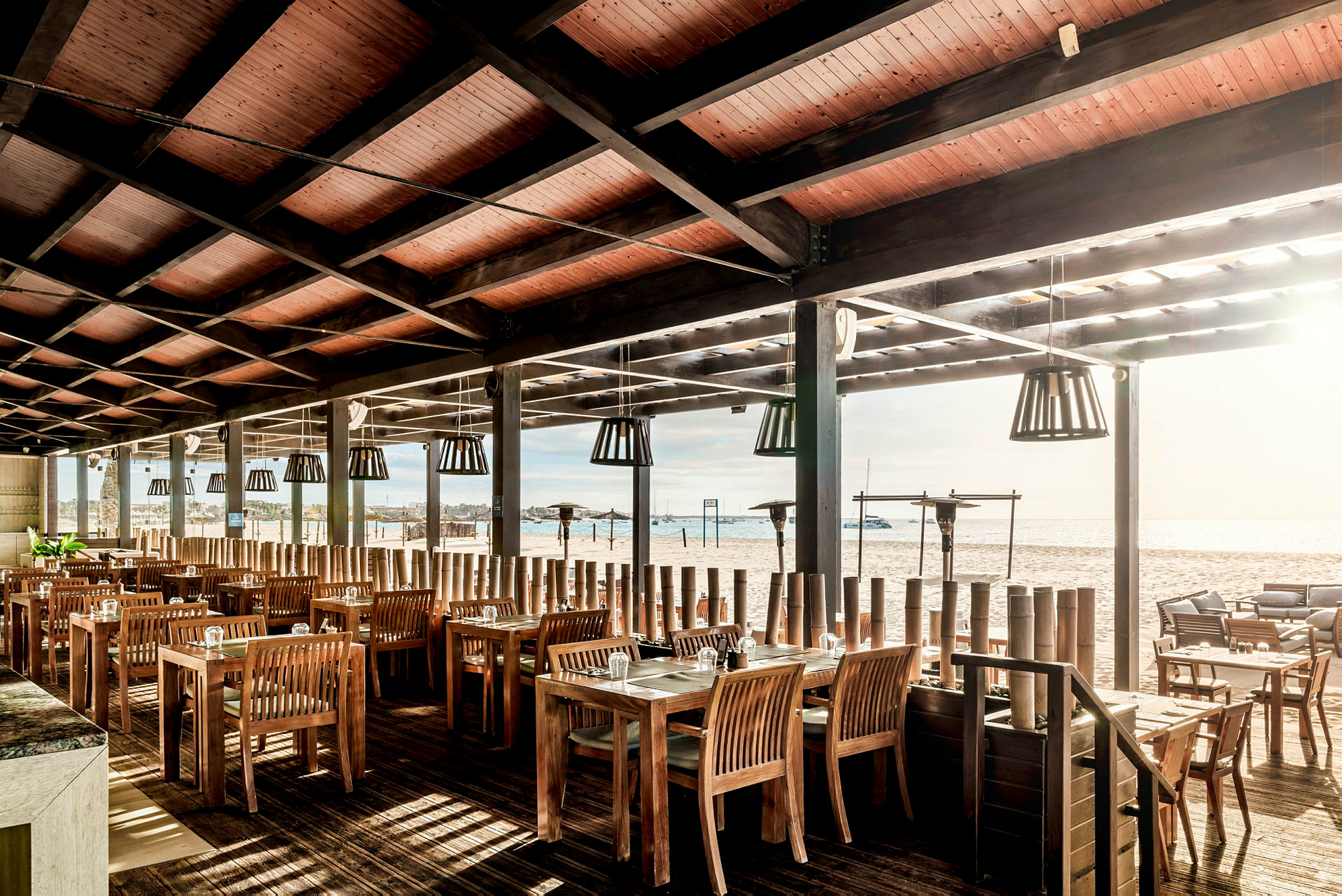 Restaurang med trämöbler och tak, vid stranden med utsikt över vattnet. (AI-genererad text)