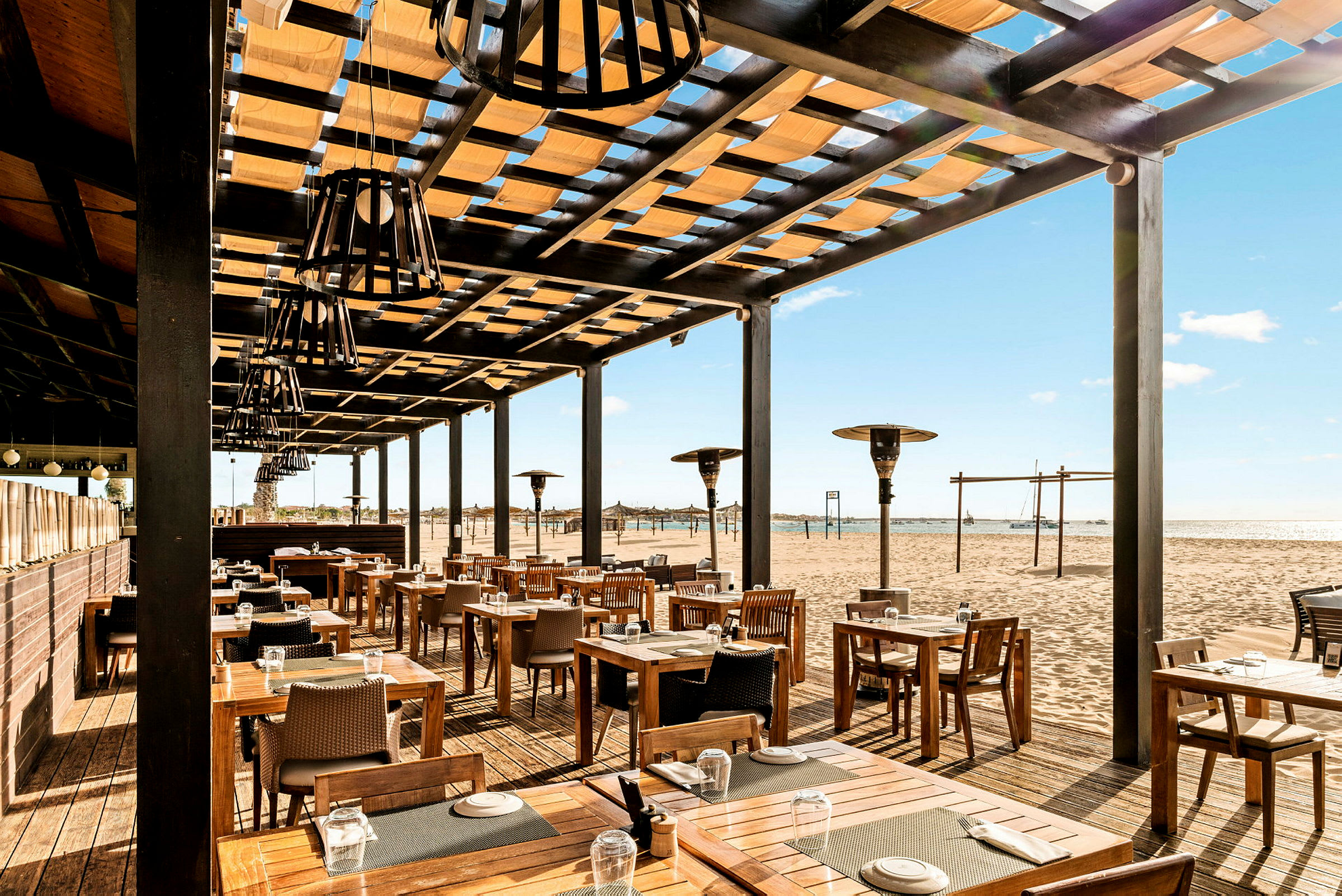 Utomhusrestaurang med träbord och stolar, belägen vid en sandstrand. (AI-genererad text)