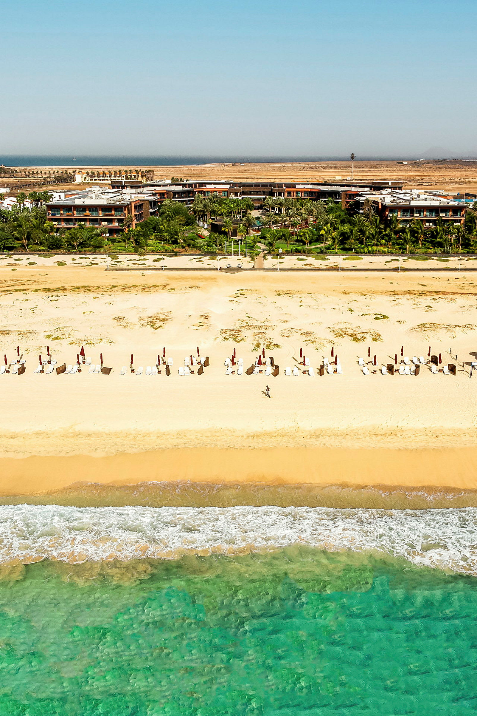 Bild av en strand med solstolar och parasoller, på avstånd syns gröna träd och byggnader. (AI-genererad text)