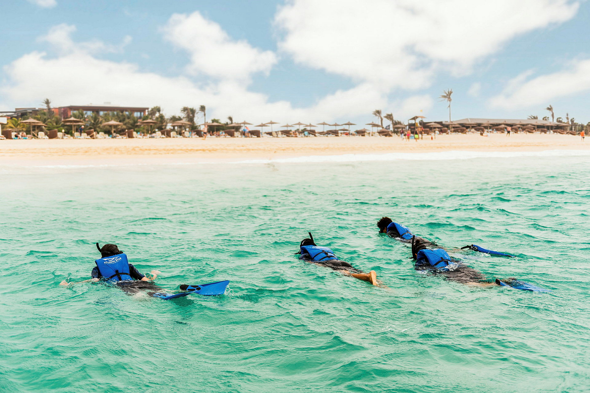Tre personer snorklar i havet nära en sandstrand med parasoller och byggnader i bakgrunden. (AI-genererad text)