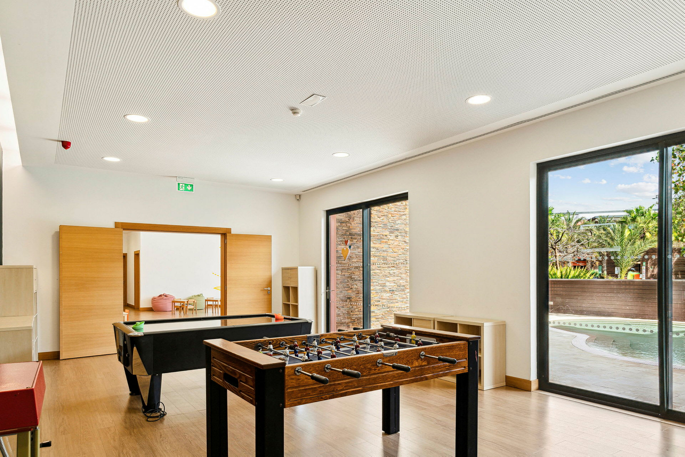 Ett spelrum med ett foosbollbord, ett biljardbord och en glasyta med utsikt mot en pool. (AI-genererad text)