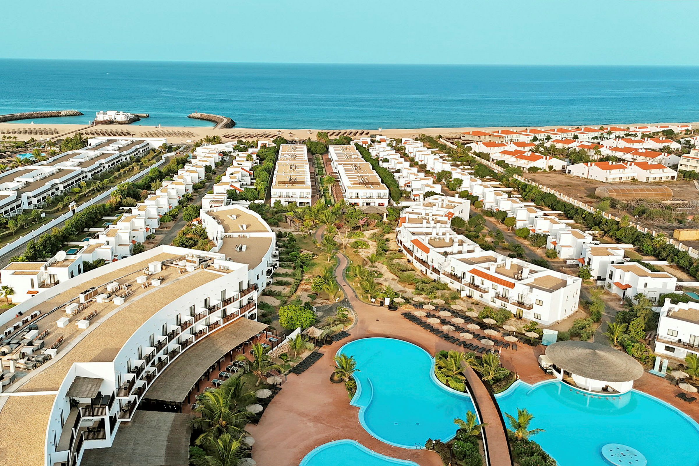 Flygbild av ett hotellområde med pooler, byggnader och strand i bakgrunden. (AI-genererad text)