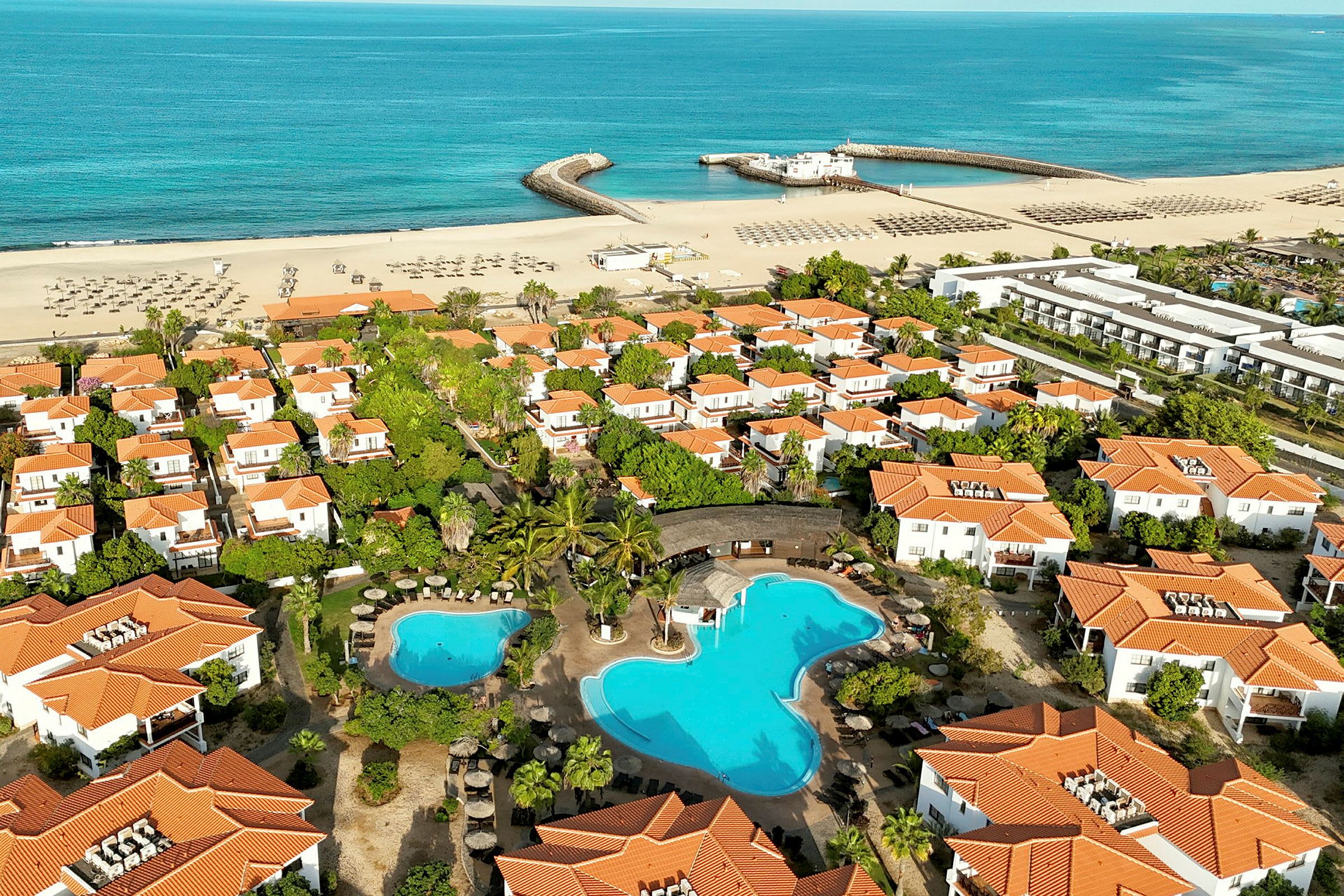 Flygvy över ett semesterområde med vita hus med orangea tak, swimmingpool och en sandstrand vid havet. (AI-genererad text)