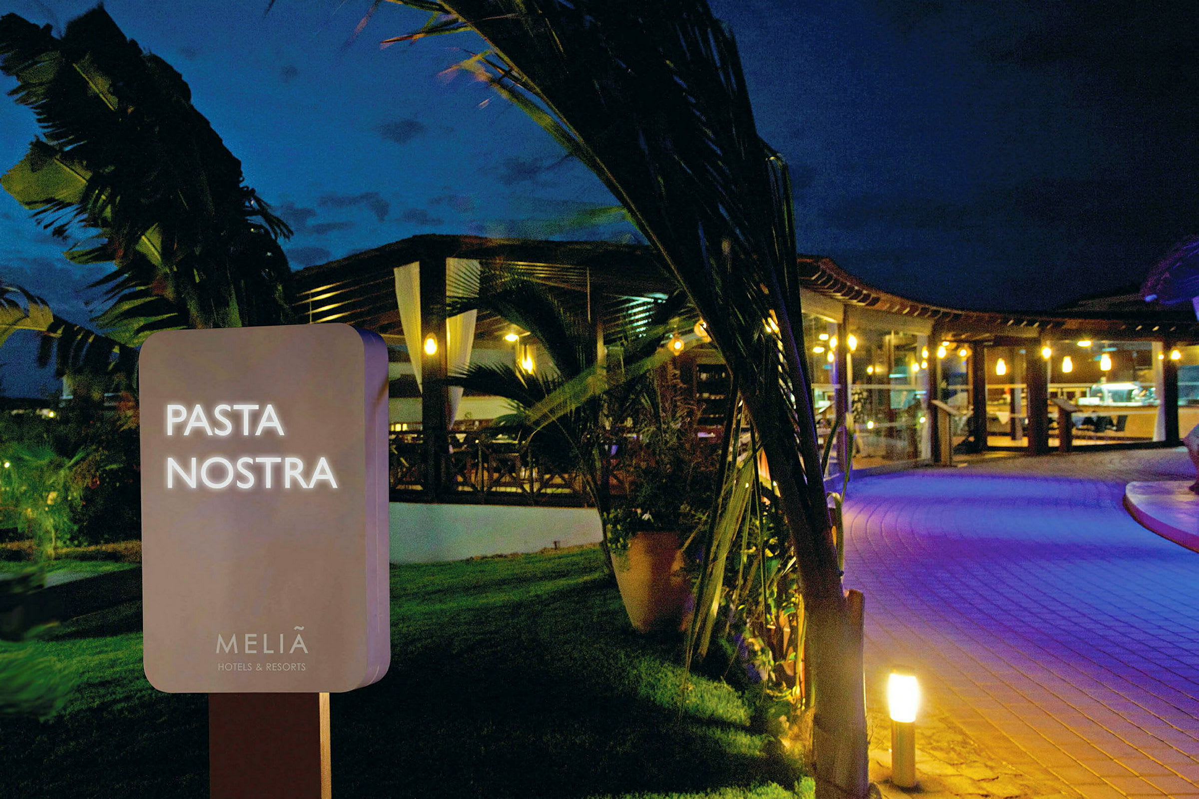 Skylt med texten 'Pasta Nostra' framför en upplyst restaurang på kvällen. (AI-genererad text)