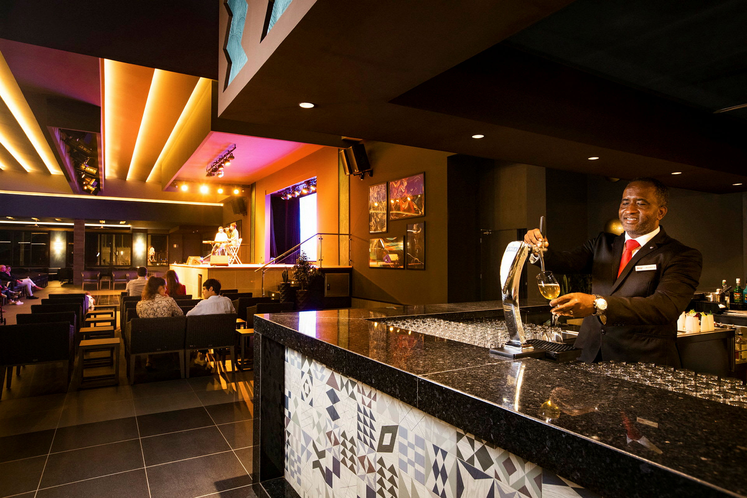 En elegant bar med en bartender som serverar dryck samt en upplyst scen i bakgrunden med sittande gäster. (AI-genererad text)
