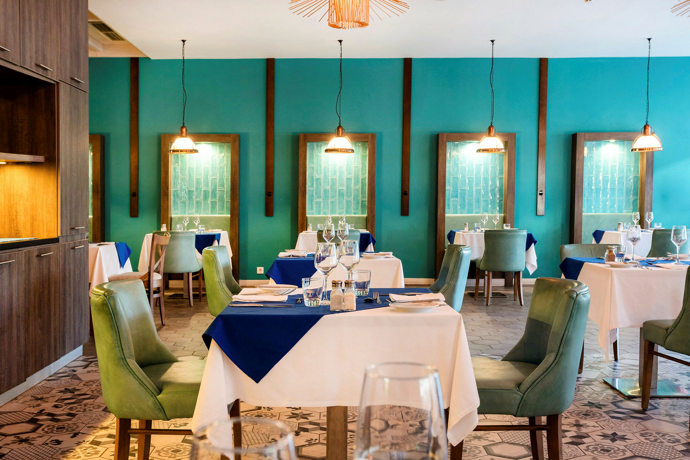 Ett elegant restauranginteriör med dukade bord och gröna stolar, omgiven av turkosa väggar och dekorativa hänglampor. (AI-genererad text)