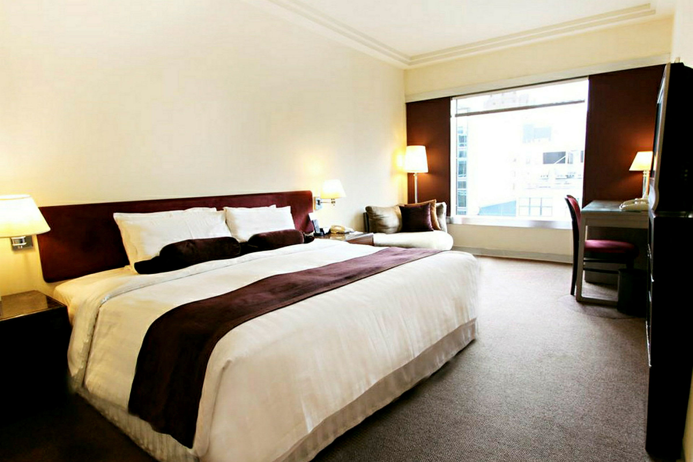 Hotellrum med en stor säng, ett skrivbord och en fåtölj bredvid ett stort fönster. (AI-genererad text)