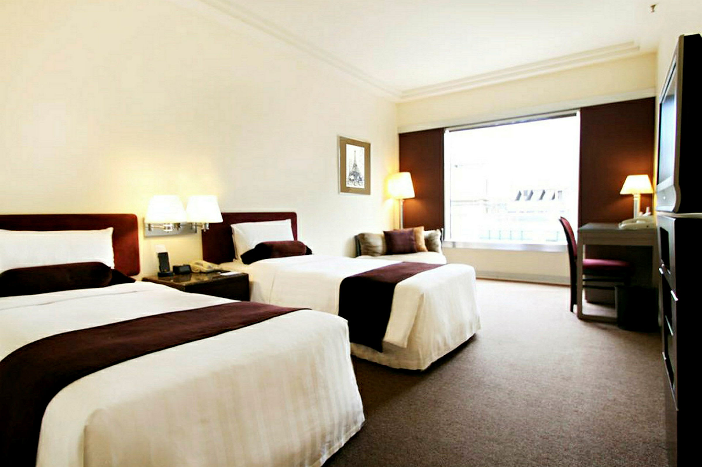 Ett hotellrum med två sängar utrustade med vita lakan och bruna överkast. Det finns ett skrivbord med stol samt fönster mot utsidan. (AI-genererad text)