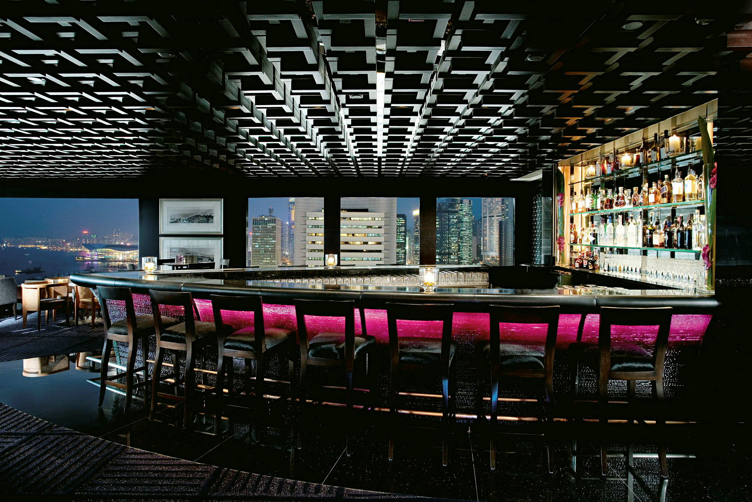 Bild av en lyxig bar med hög ryggstolar vid bardisken. Barens belysning är stämningsfull med en neonrosa ton och stora fönster visar utsikt över stadens skyline. (AI-genererad text)
