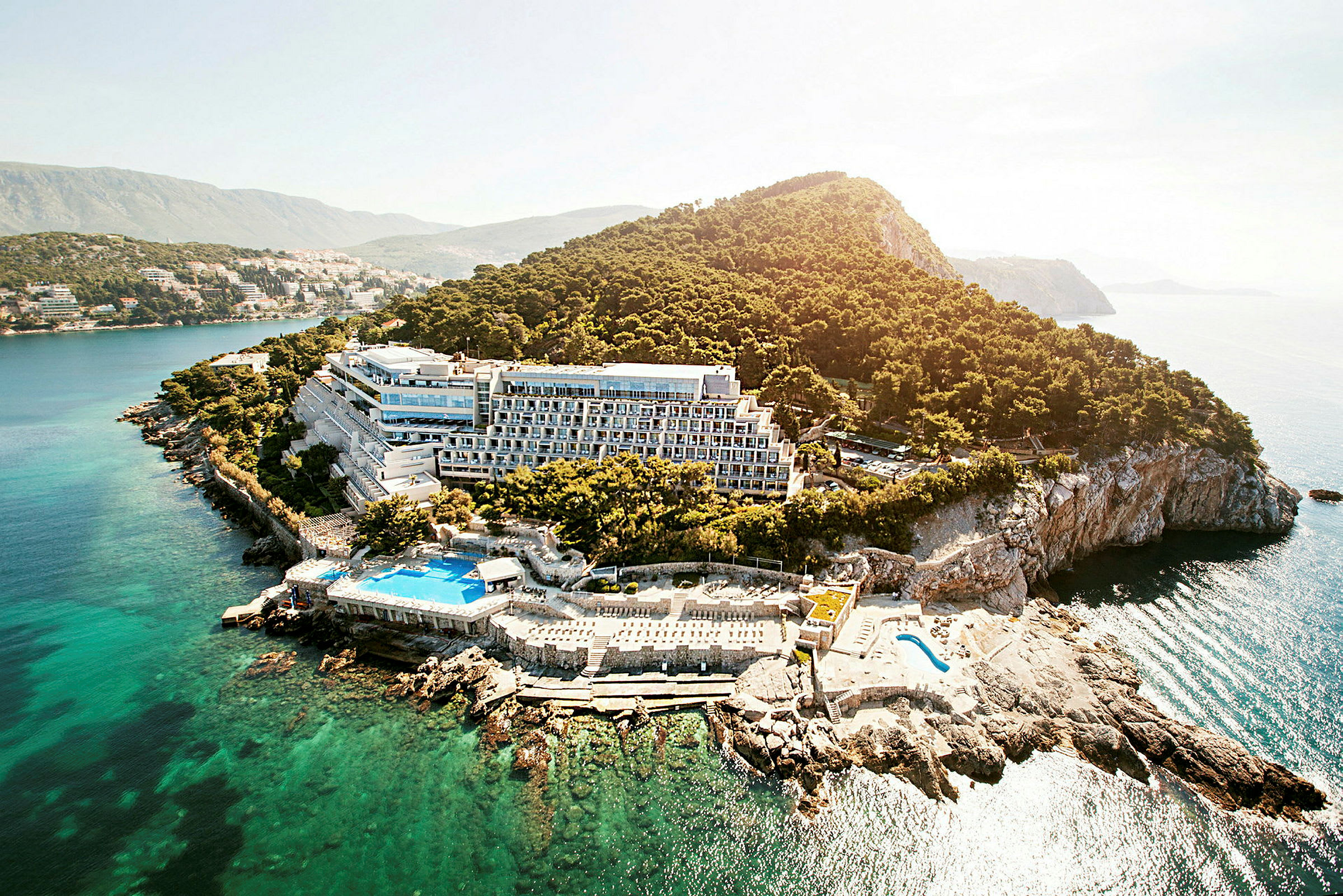 Flygfoto av en hotellbyggnad belägen vid en klippig kust omgiven av träd och hav. (AI-genererad text)
