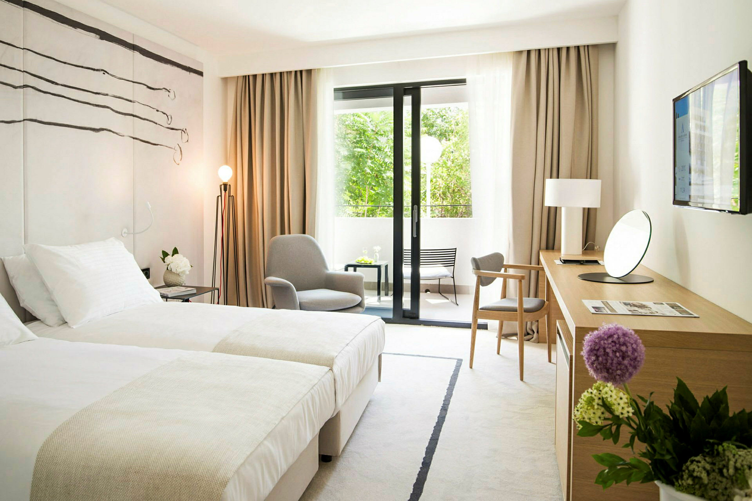 Ett modernt hotellrum med två sängar, en fåtölj, ett skrivbord med en spegel och en TV på väggen. Det finns stora fönsterdörrar som leder till en utomhusbalkong med utsikt mot grönska. (AI-genererad text)