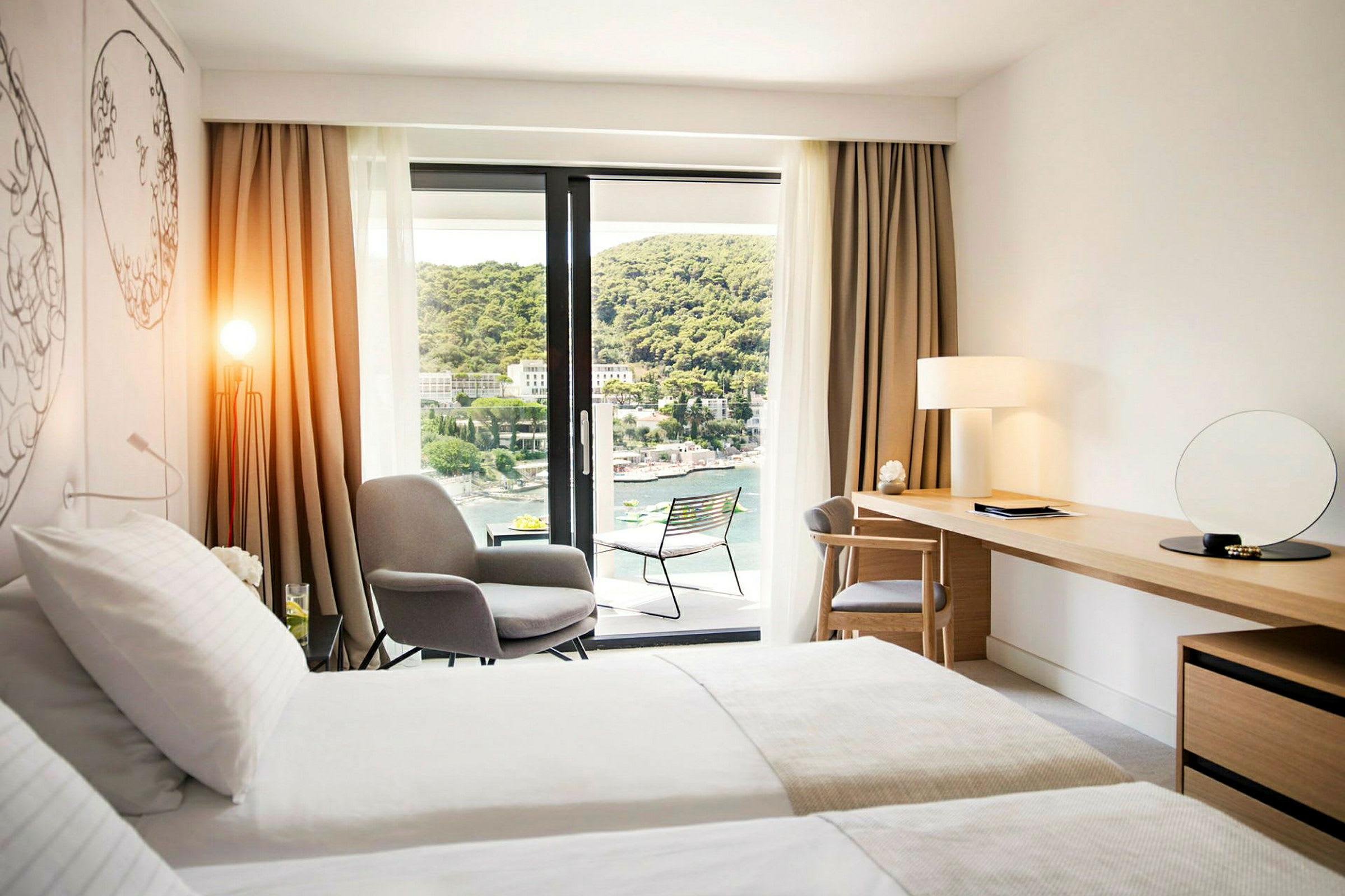 Modernt hotellrum med dubbelsäng, sittplats, skrivbord och balkong med utsikt över en grönskande sluttning och vatten. (AI-genererad text)