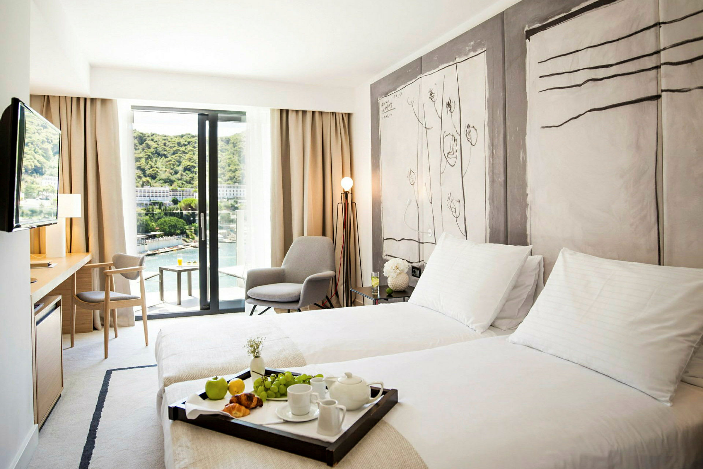 Ett hotellrum med två enkelsängar, en sittgrupp och en balkong med utsikt över en grönskande omgivning. (AI-genererad text)