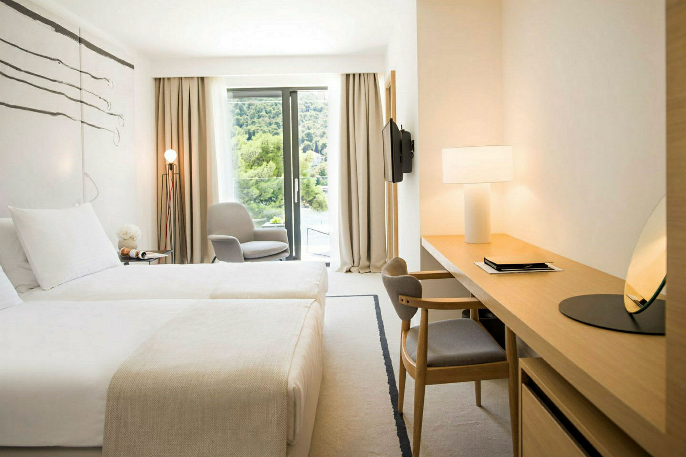 Ett ljust hotellrum med två sängar, en skrivbordshörna, en stol vid ett fönster och en utsikt över grönska. (AI-genererad text)