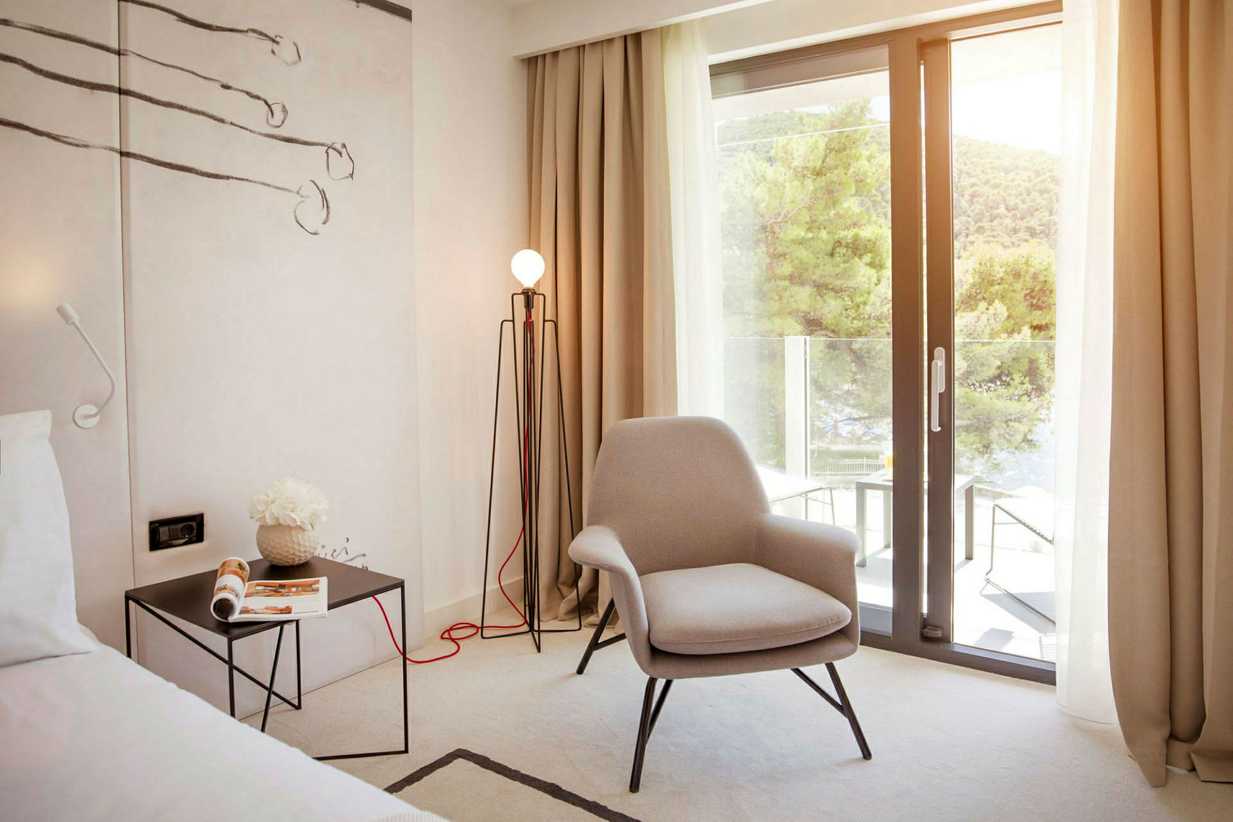 Ett hotellrum med en bekväm fåtölj, ett sidobord med en tidning och en golvlampa. En stor glasdörr öppnar upp till en balkong med utsikt över grönskande träd. (AI-genererad text)