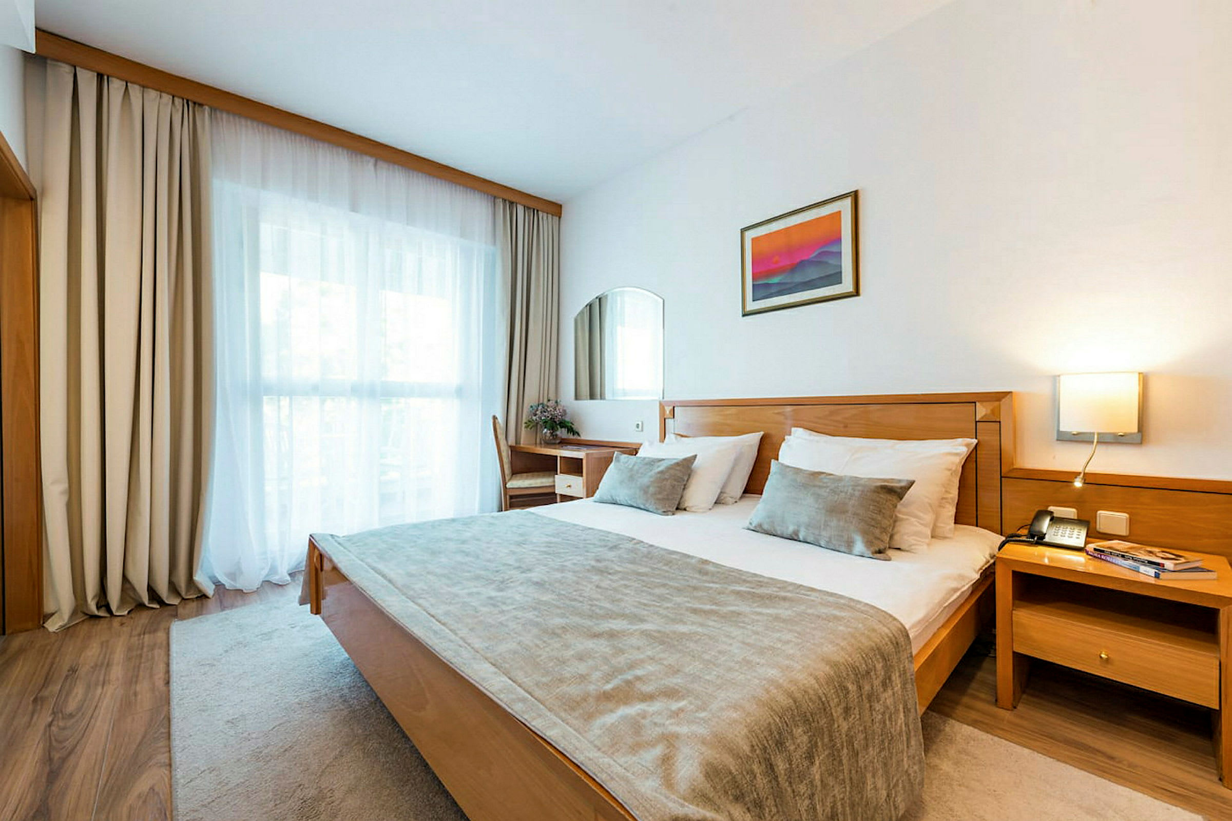 Ett hotellrum med en dubbelsäng, en skrivbordsstol, ett fönster med gardiner och en väggprydnad. (AI-genererad text)