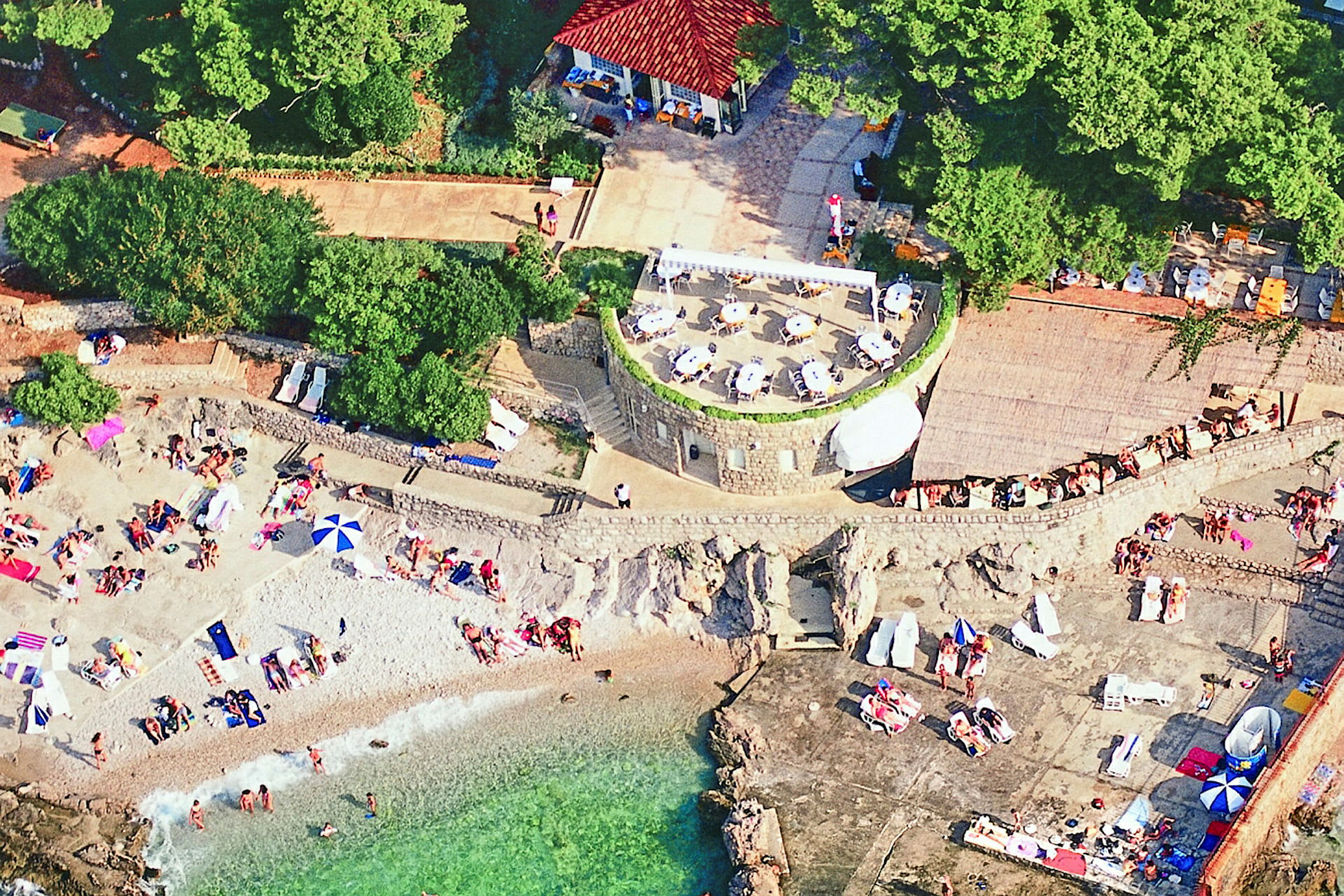 Flygfoto över en strand med solande människor och en byggnad med uteservering i närheten. (AI-genererad text)