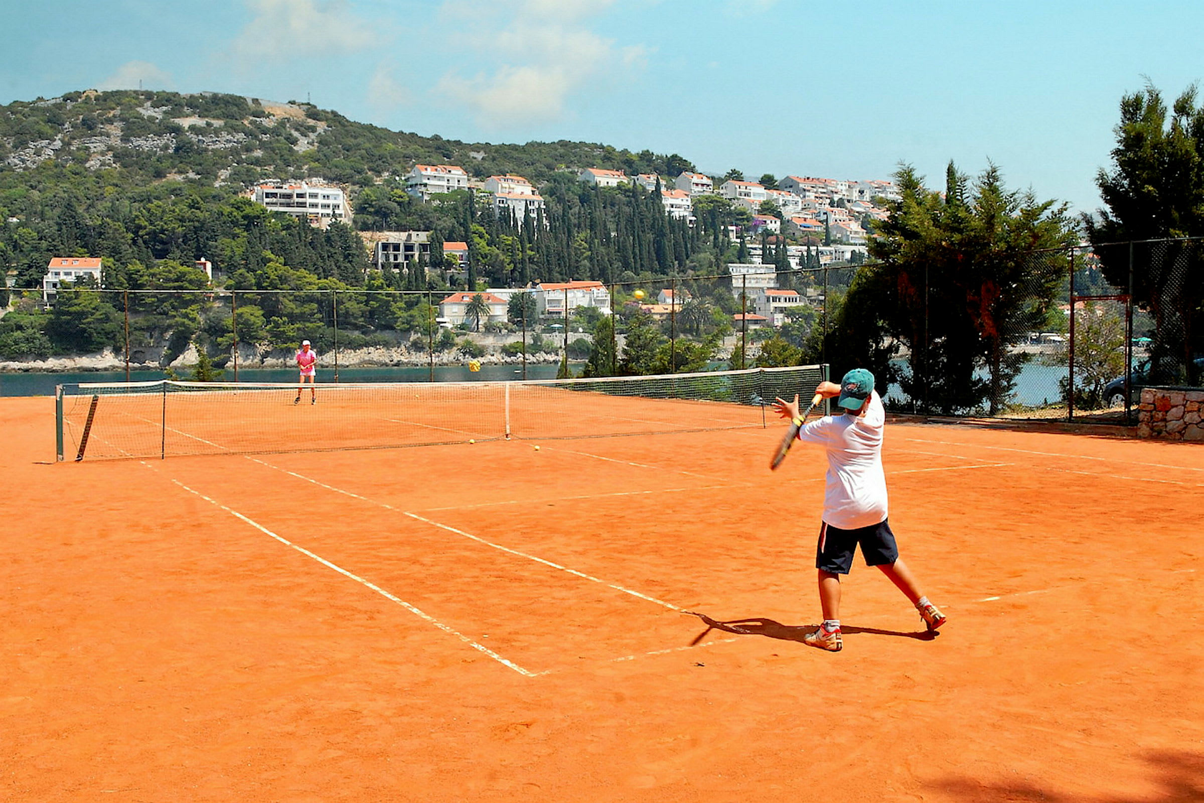 Två personer spelar tennis på en utomhusplan med grusbeläggning. Bakom tennisplanen syns en intilliggande kulle med byggnader och grönska. (AI-genererad text)