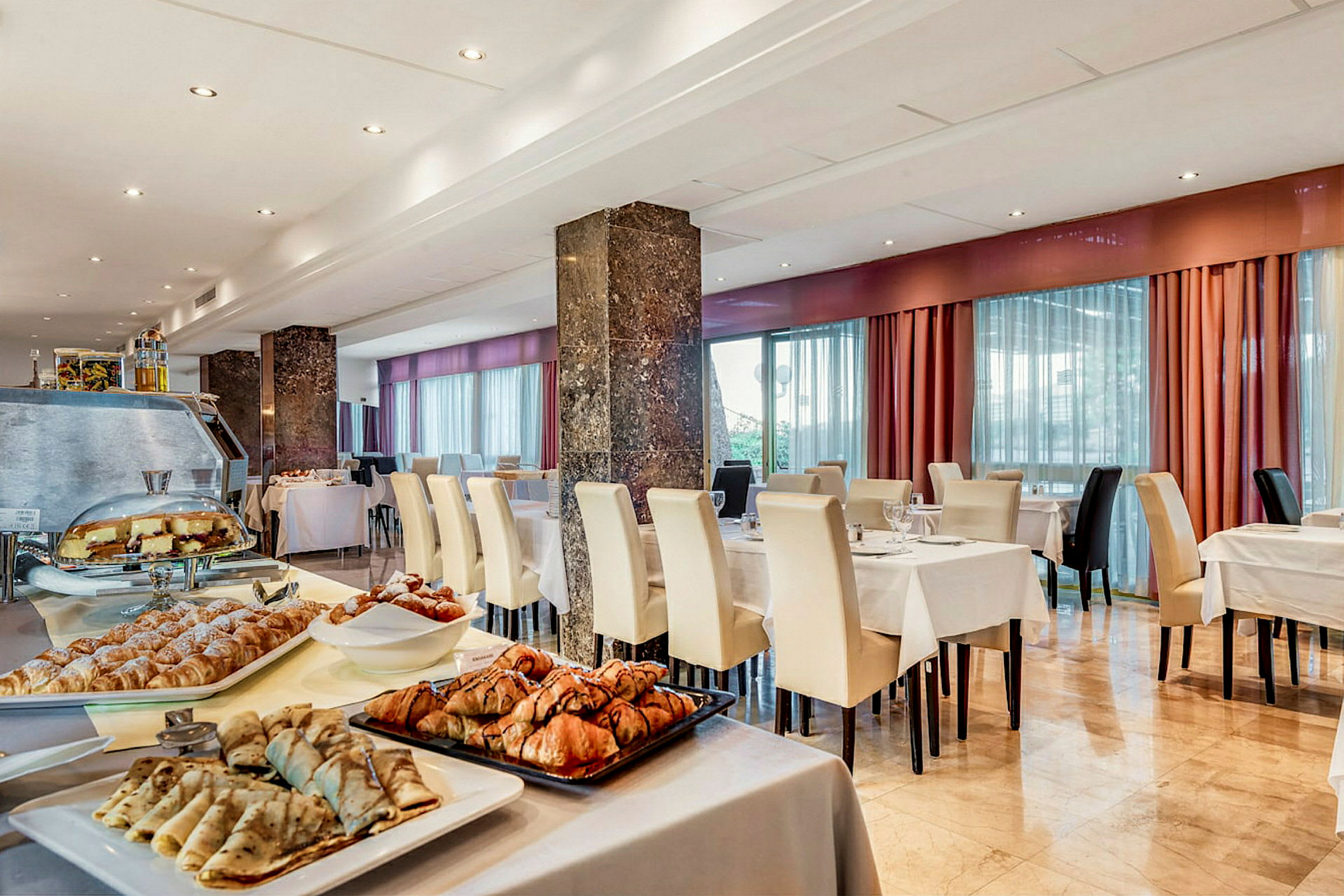 En hotellrestaurang med dukade bord och vita stolar, samt ett frukostbuffébord med olika bakverk och pannkakor. (AI-genererad text)