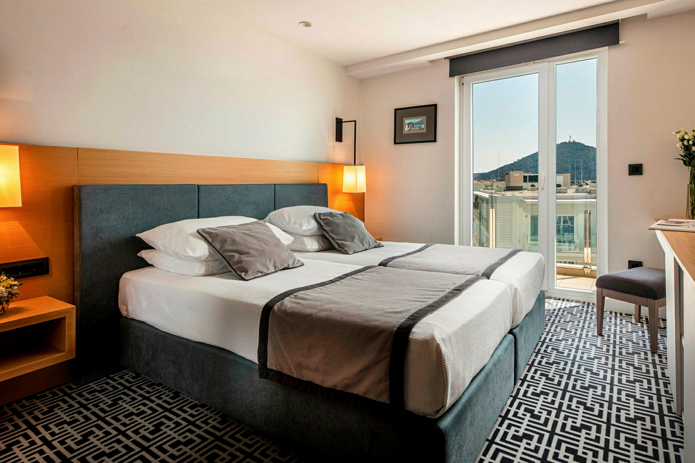 Hotellrum med dubbelsäng, ljus inredning och balkong med utsikt över stad och berg. (AI-genererad text)