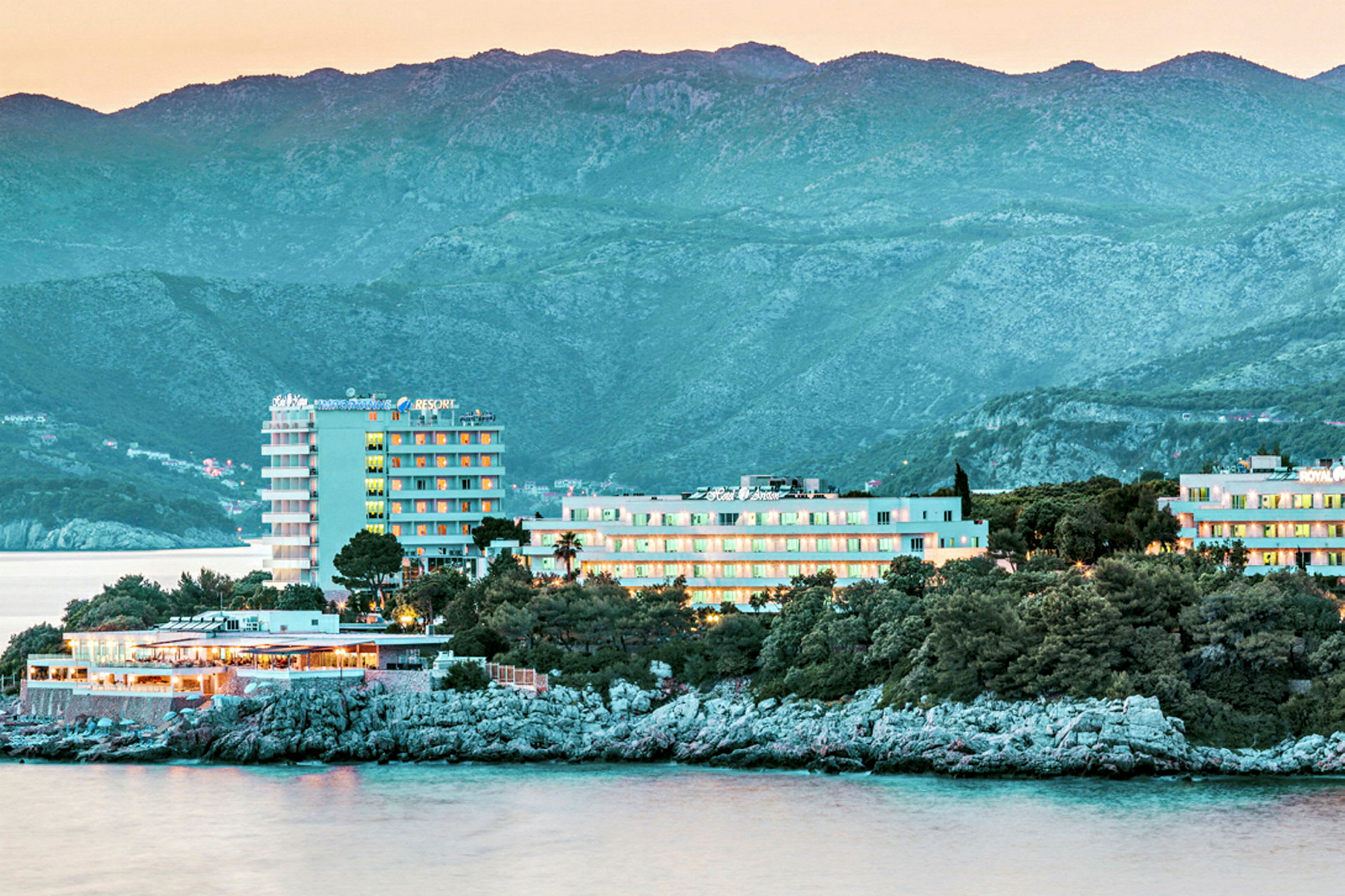 Ett kustnära hotell med utsikt över havet, omgivet av träd och grönska, med berg i bakgrunden. (AI-genererad text)