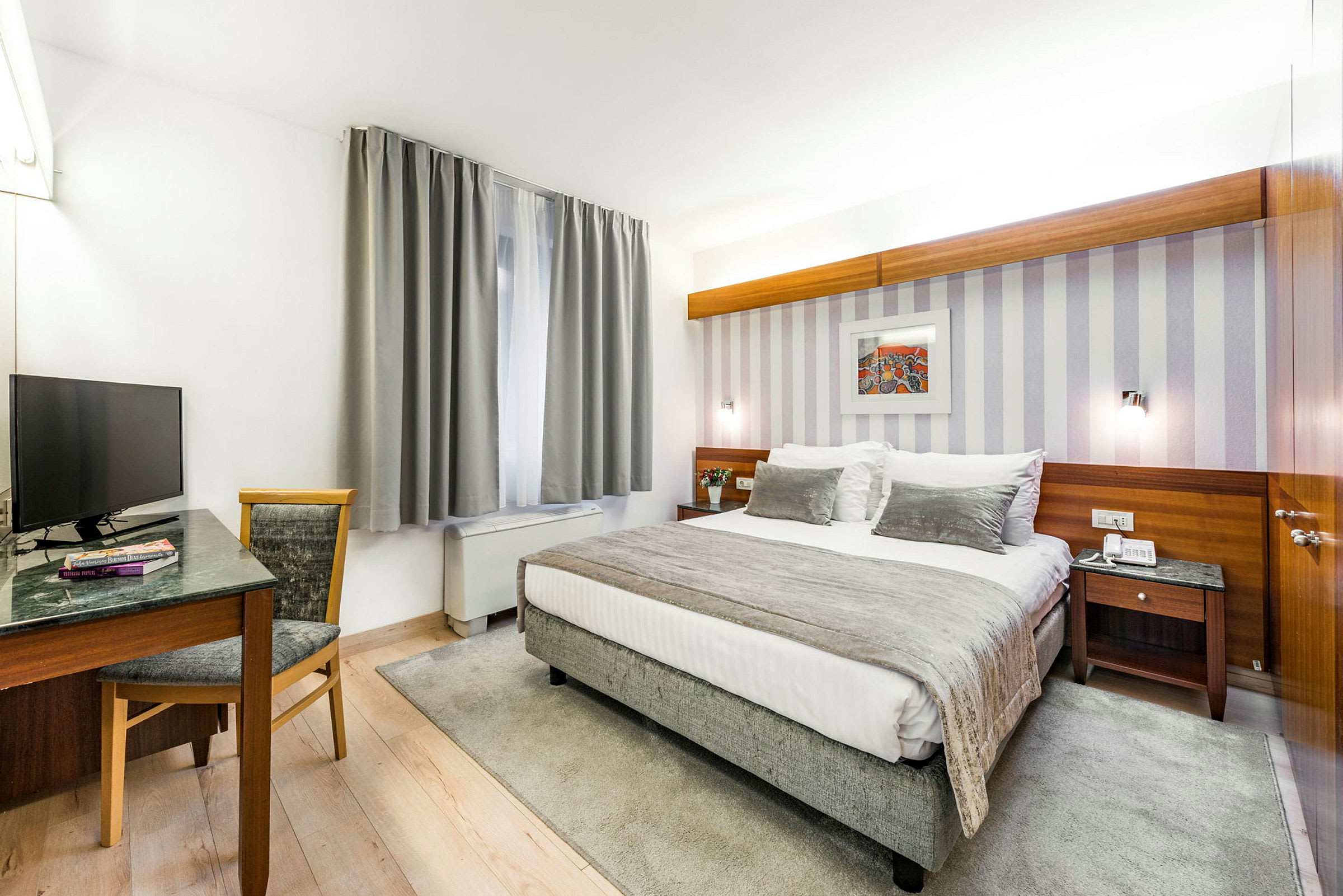 Ett hotellrum med en dubbelsäng och ett skrivbord bredvid ett fönster med grå gardiner. (AI-genererad text)