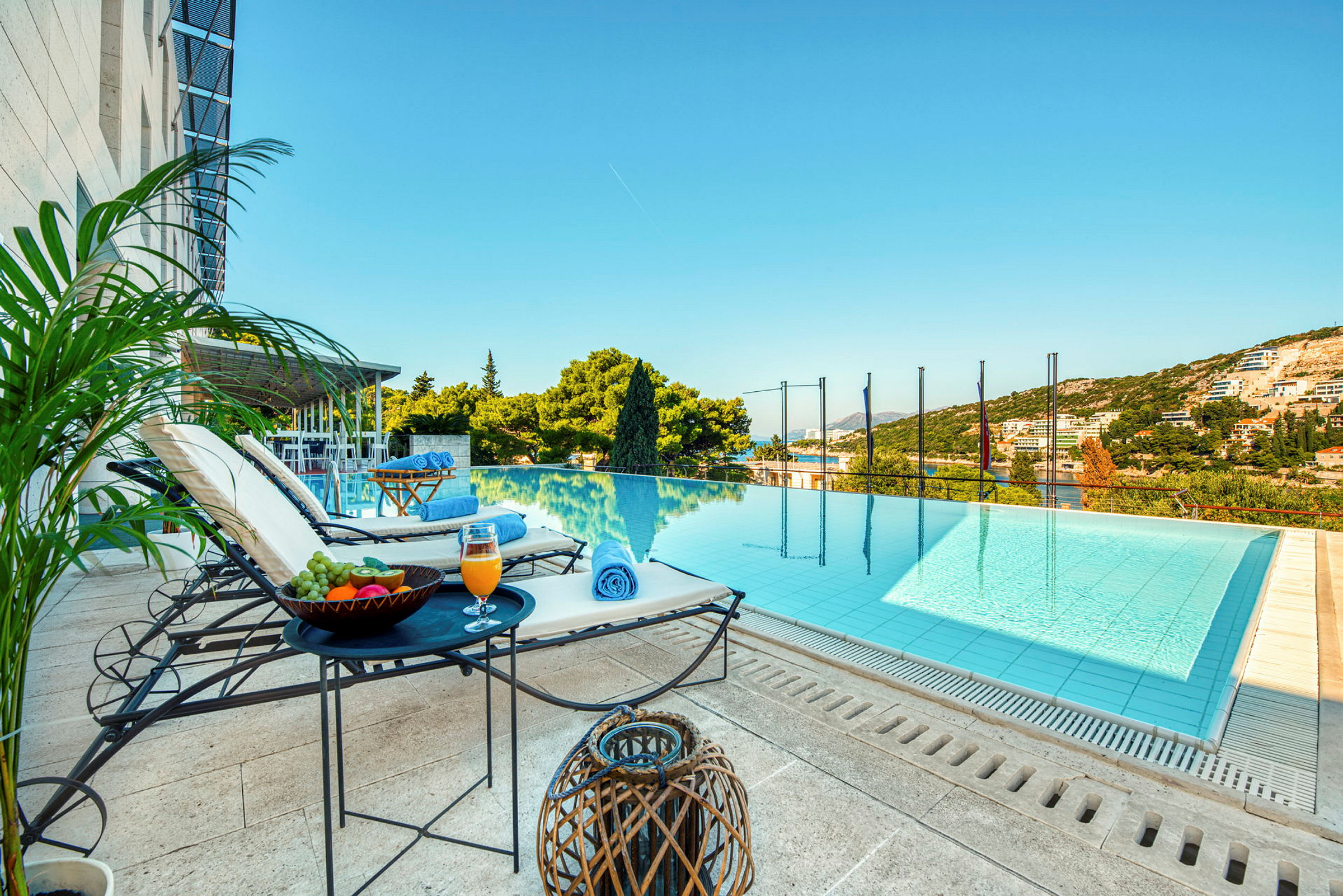 En utsikt mot en infinitypool med solstolar, bord med frukt och dryck intill sig, samt gröna omgivningar och himmel i bakgrunden. (AI-genererad text)