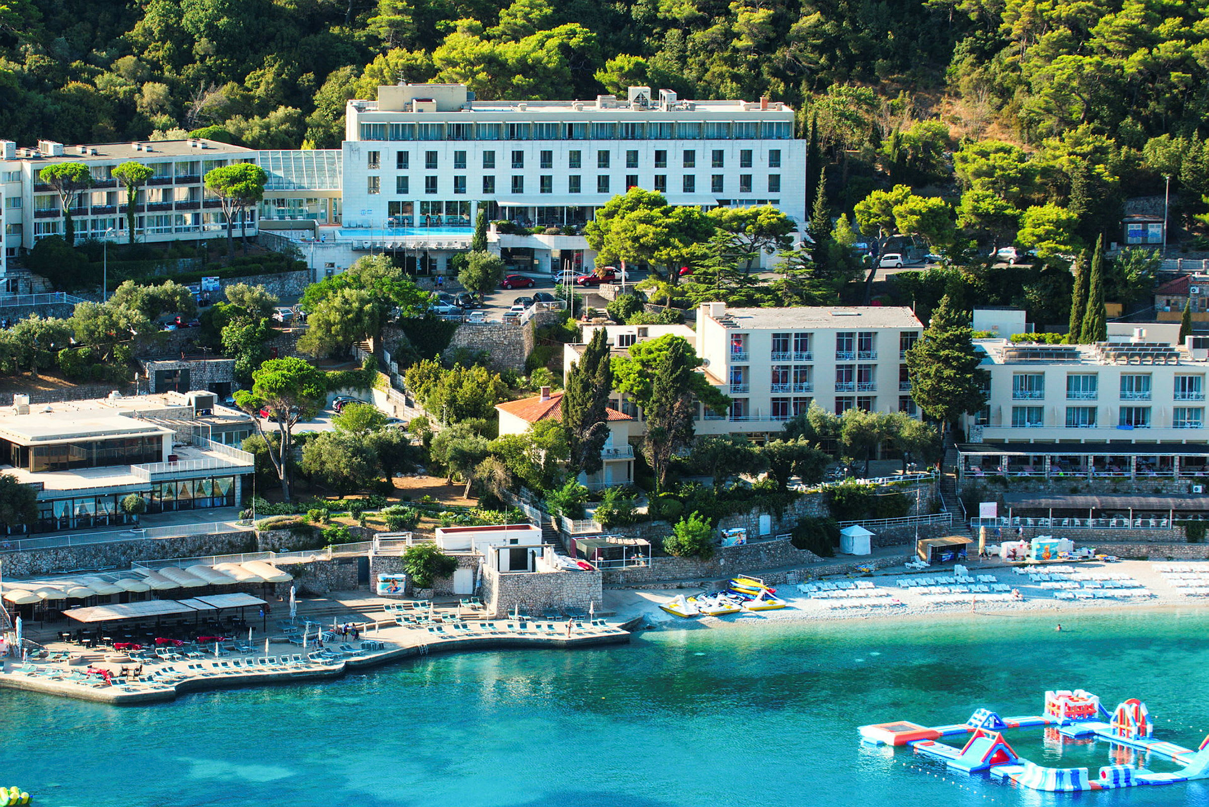 En vy över en hotellbyggnad vid en kust med solstolar på stranden och en flytande lekplats i vattnet. (AI-genererad text)