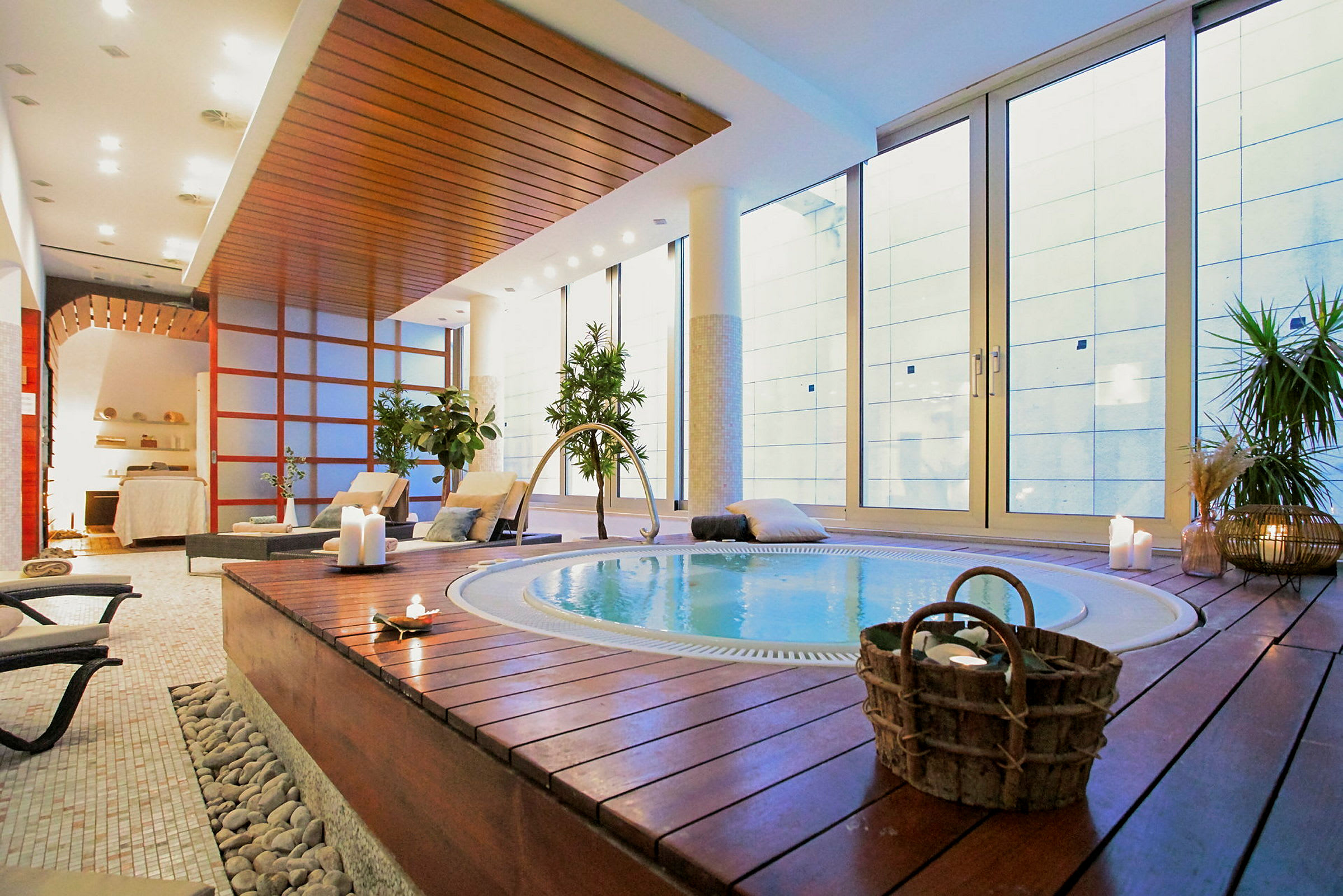 Ett elegant spa med en jacuzzi omgiven av träpanel, stolar och levande växter. Stora glasväggar ger naturligt ljus. (AI-genererad text)