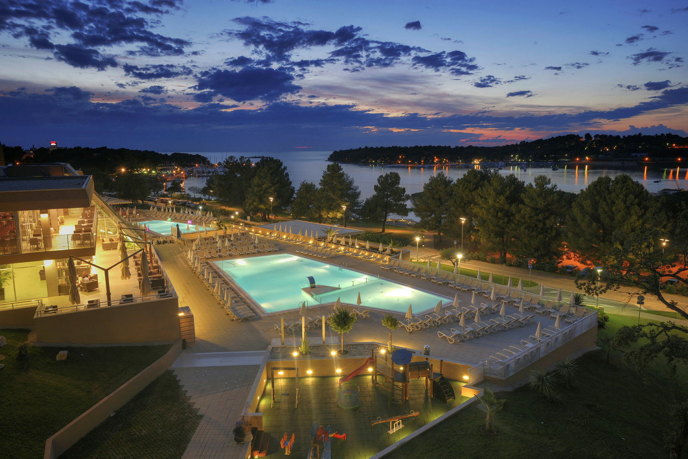 En kvällsutsikt över ett hotellområde med upplysta swimmingpooler, omgivande terrasser och träd, med en sjö i bakgrunden. (AI-genererad text)