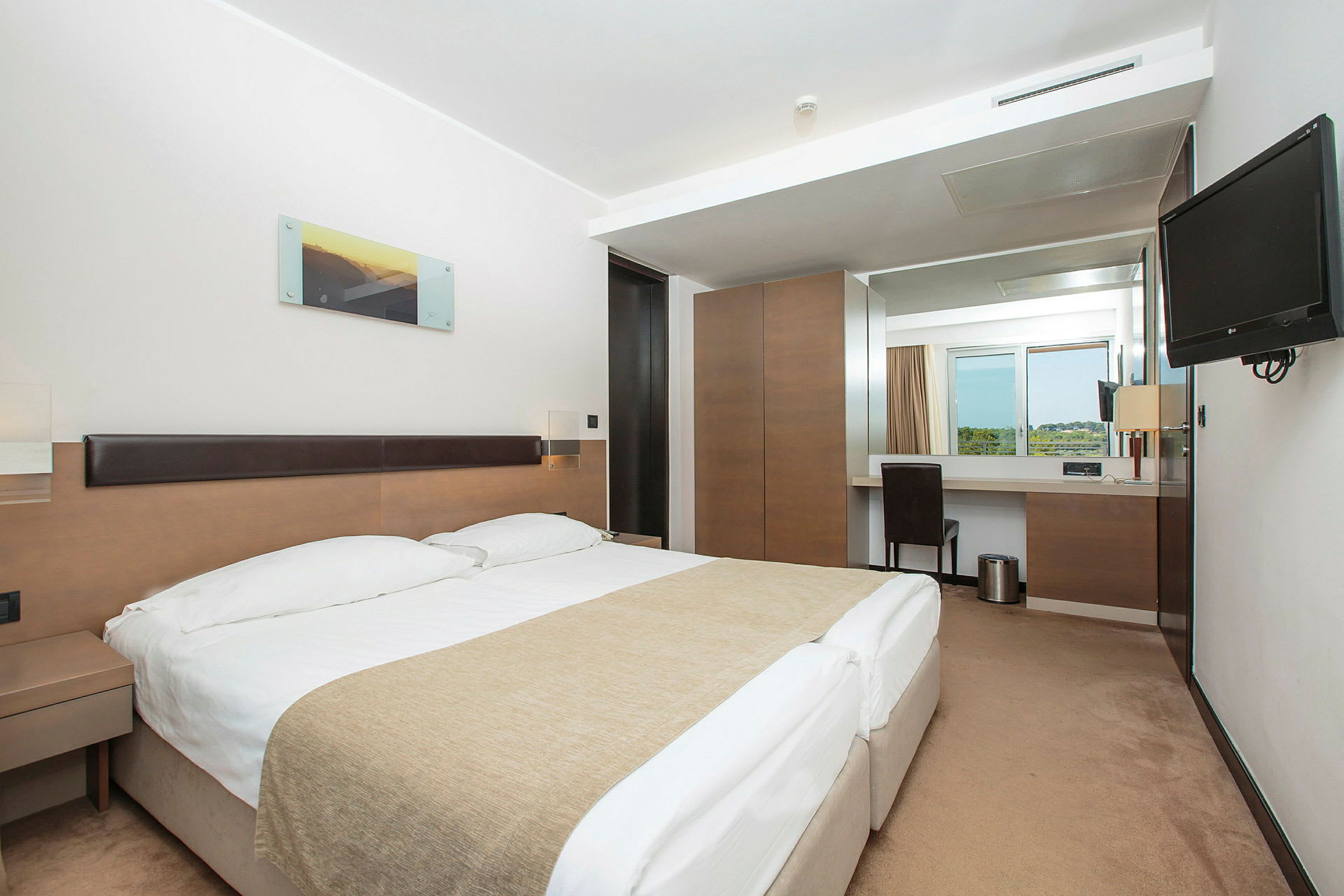 Ett hotellrum med en dubbelsäng, nattduksbord, en skrivbordshörna med stol och ett fönster med utsikt. (AI-genererad text)