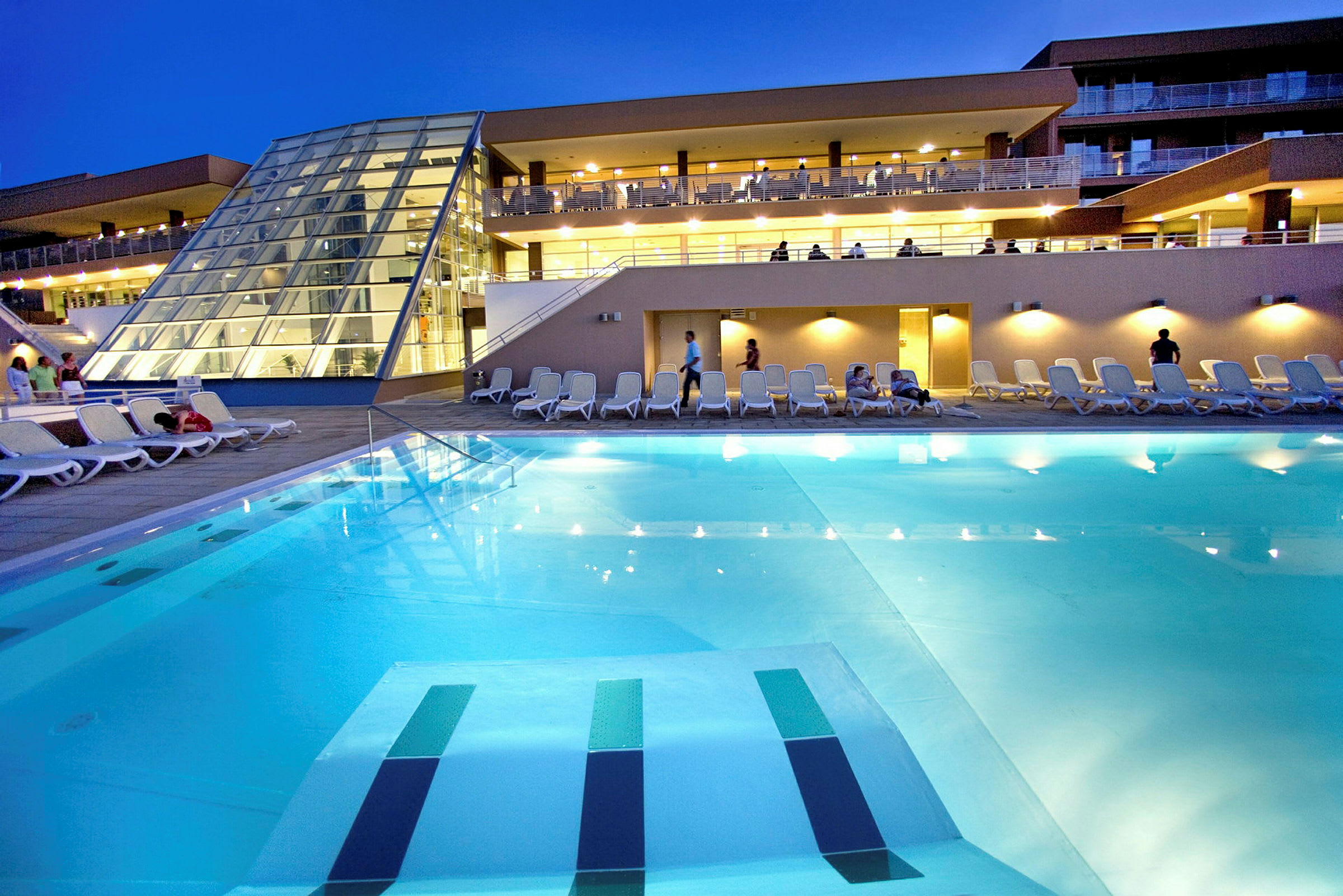 En kvällsbild av en hotellpool med solstolar runt och en modern byggnad i bakgrunden. (AI-genererad text)