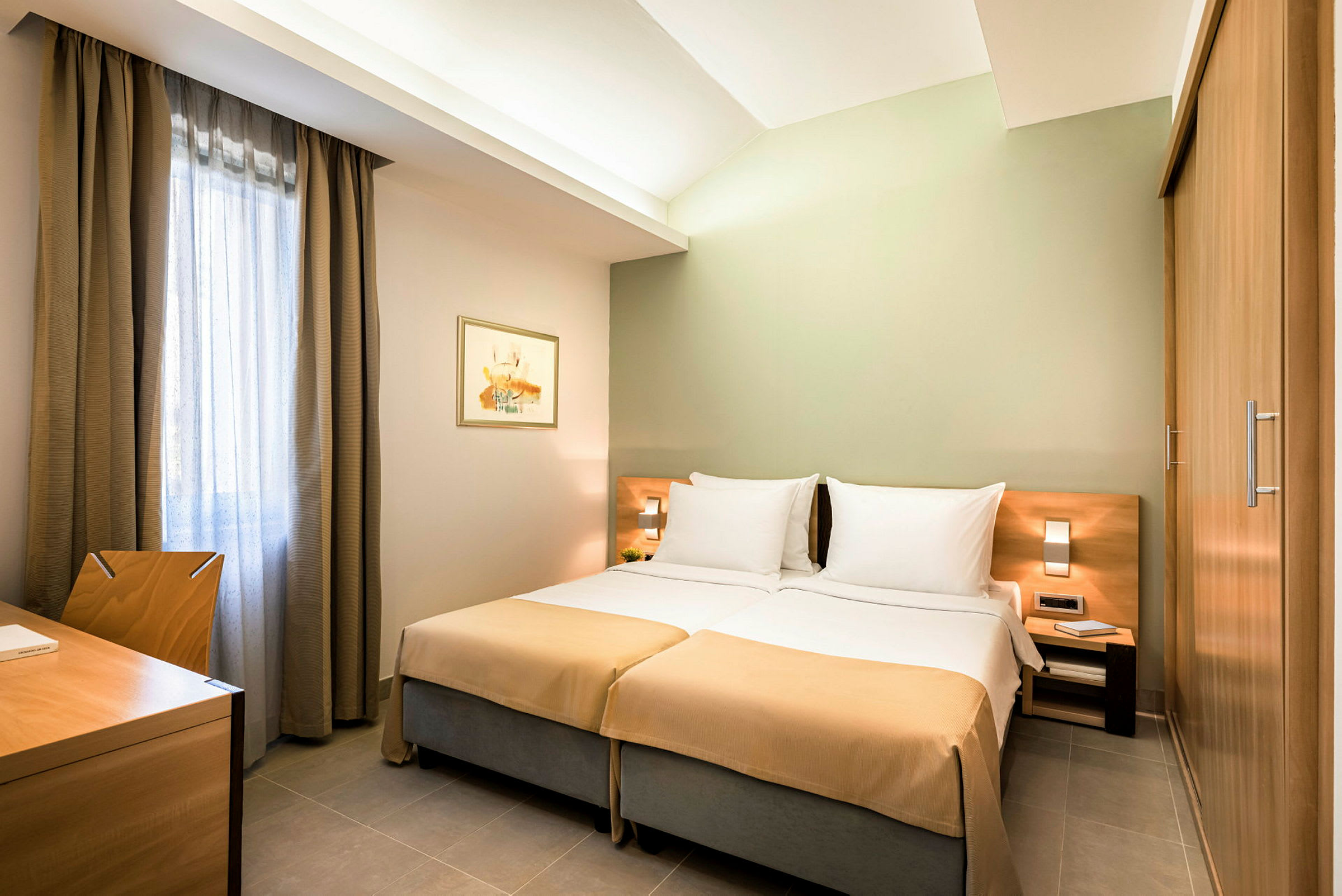 Ett hotellrum med två enkelsängar, nattduksbord, skrivbord och stol samt gardiner vid ett fönster. (AI-genererad text)