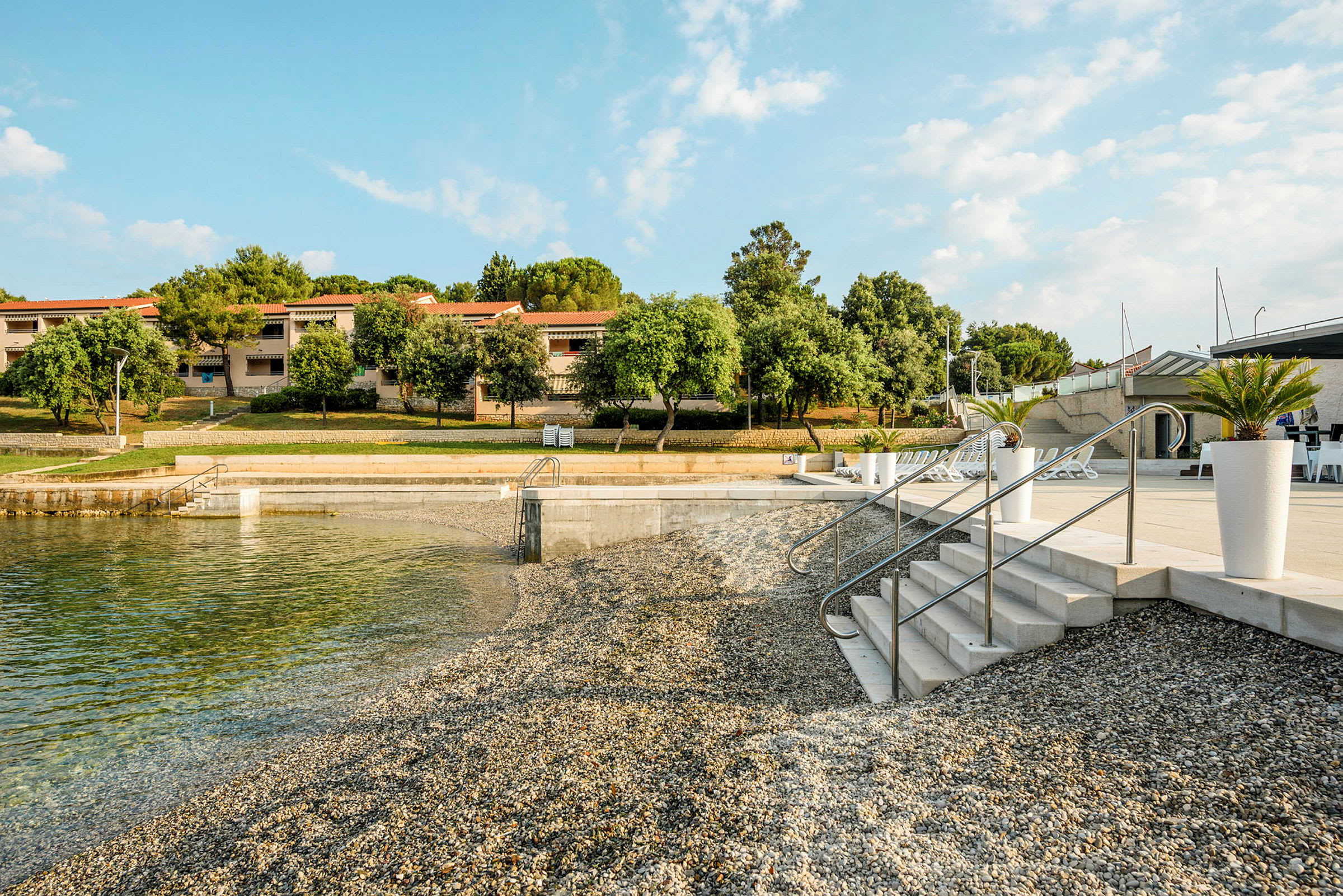 En strand med småsten och klart vatten, med trappor som leder ner till vattnet. I bakgrunden syns hus med röda tak och grönska. (AI-genererad text)