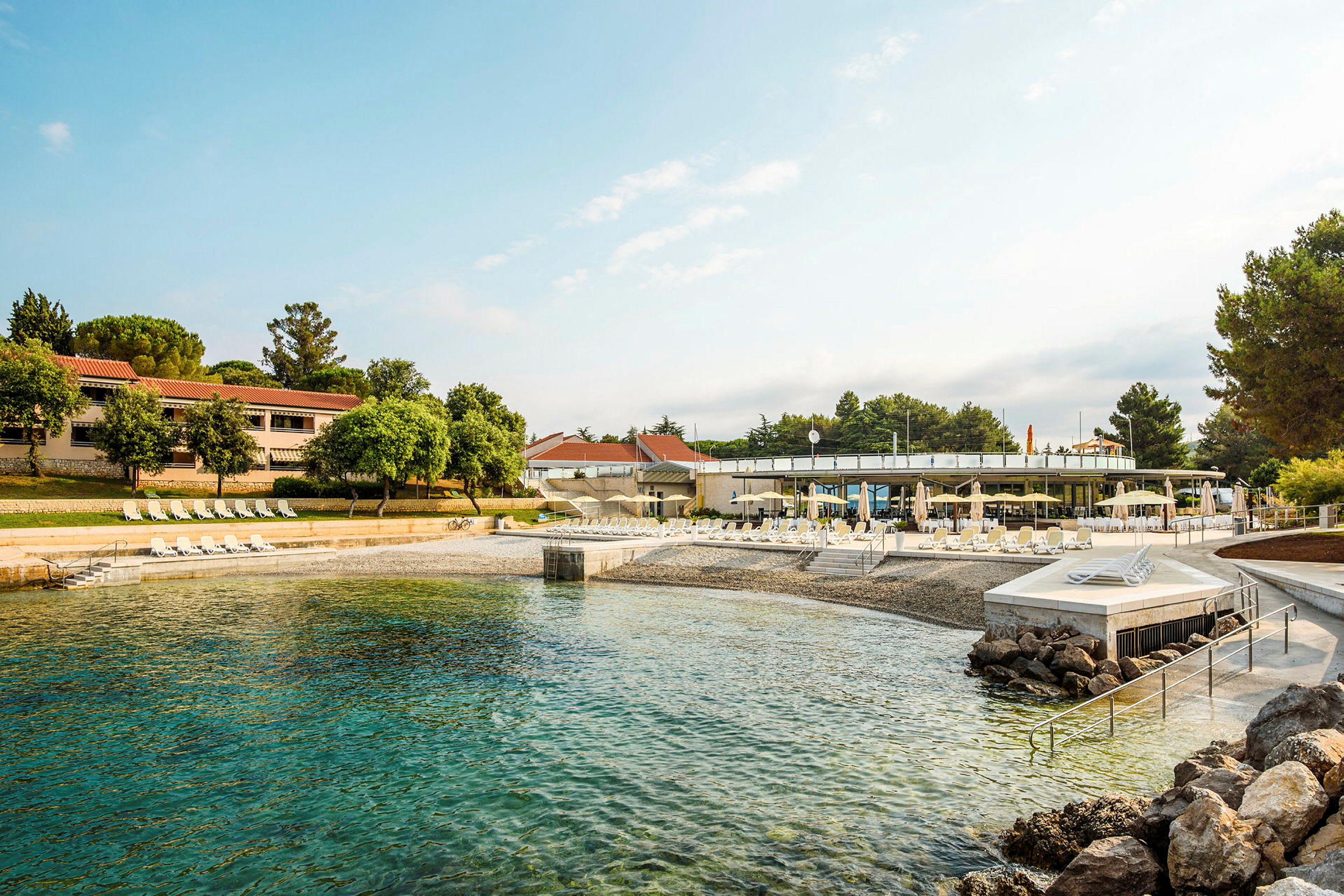 Bild av en strand med solstolar och parasoller framför byggnader och träd. (AI-genererad text)