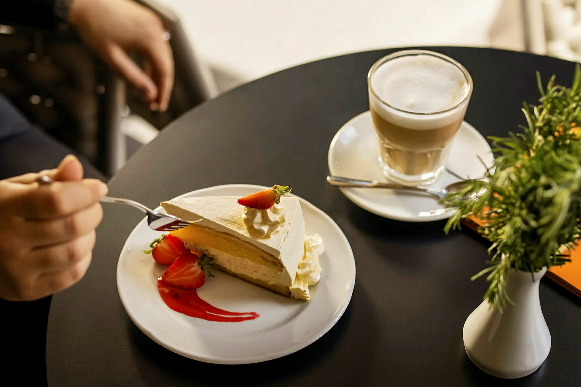 En desserttallrik med en bit tårta garnerad med jordgubbar och grädde, bredvid en kopp kaffe på ett svart bord. (AI-genererad text)